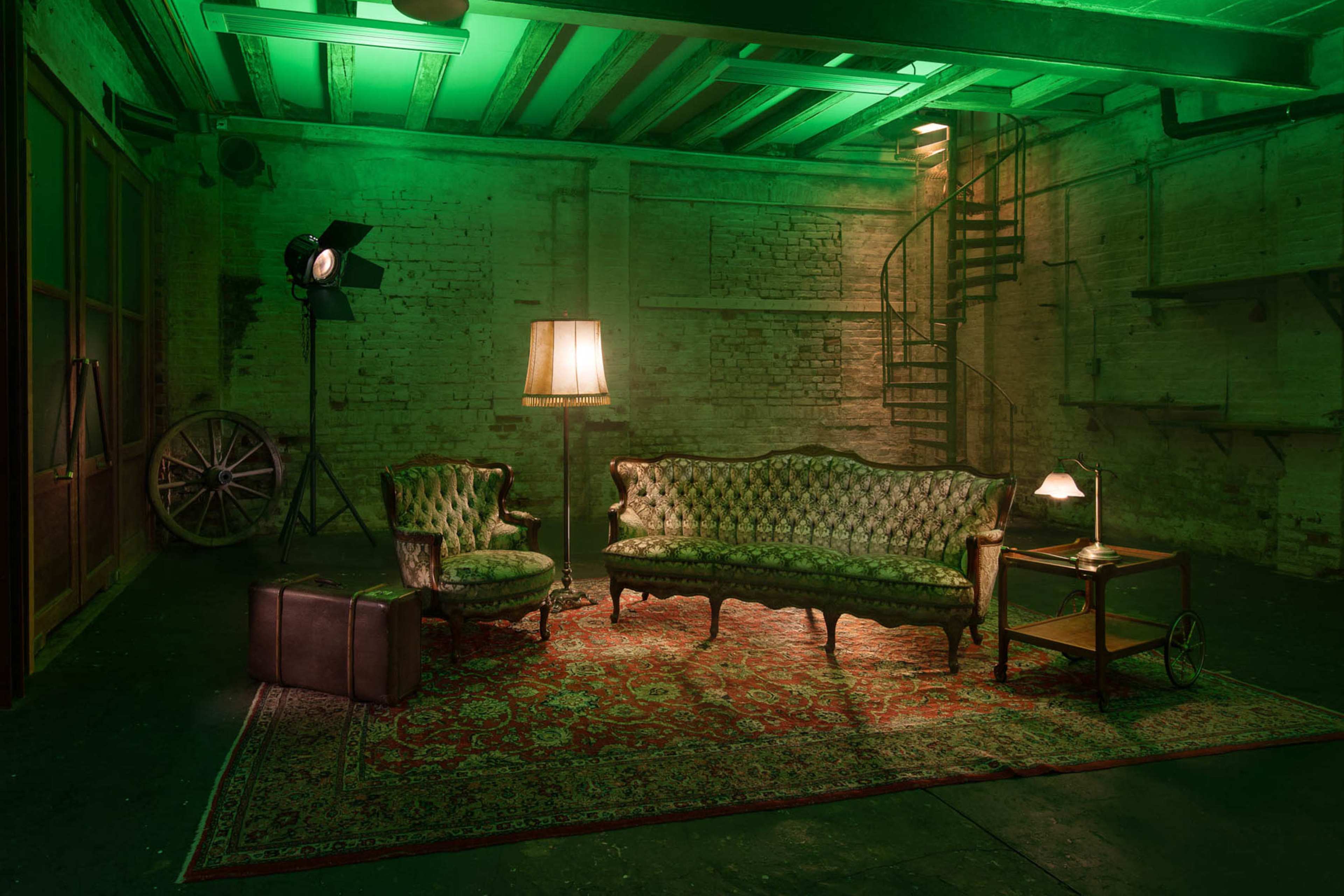 The image shows a vintage seating area with a patterned green couch and armchair, a lamp, a small table, and an old suitcase on a red patterned rug in a dimly lit room with green lighting.