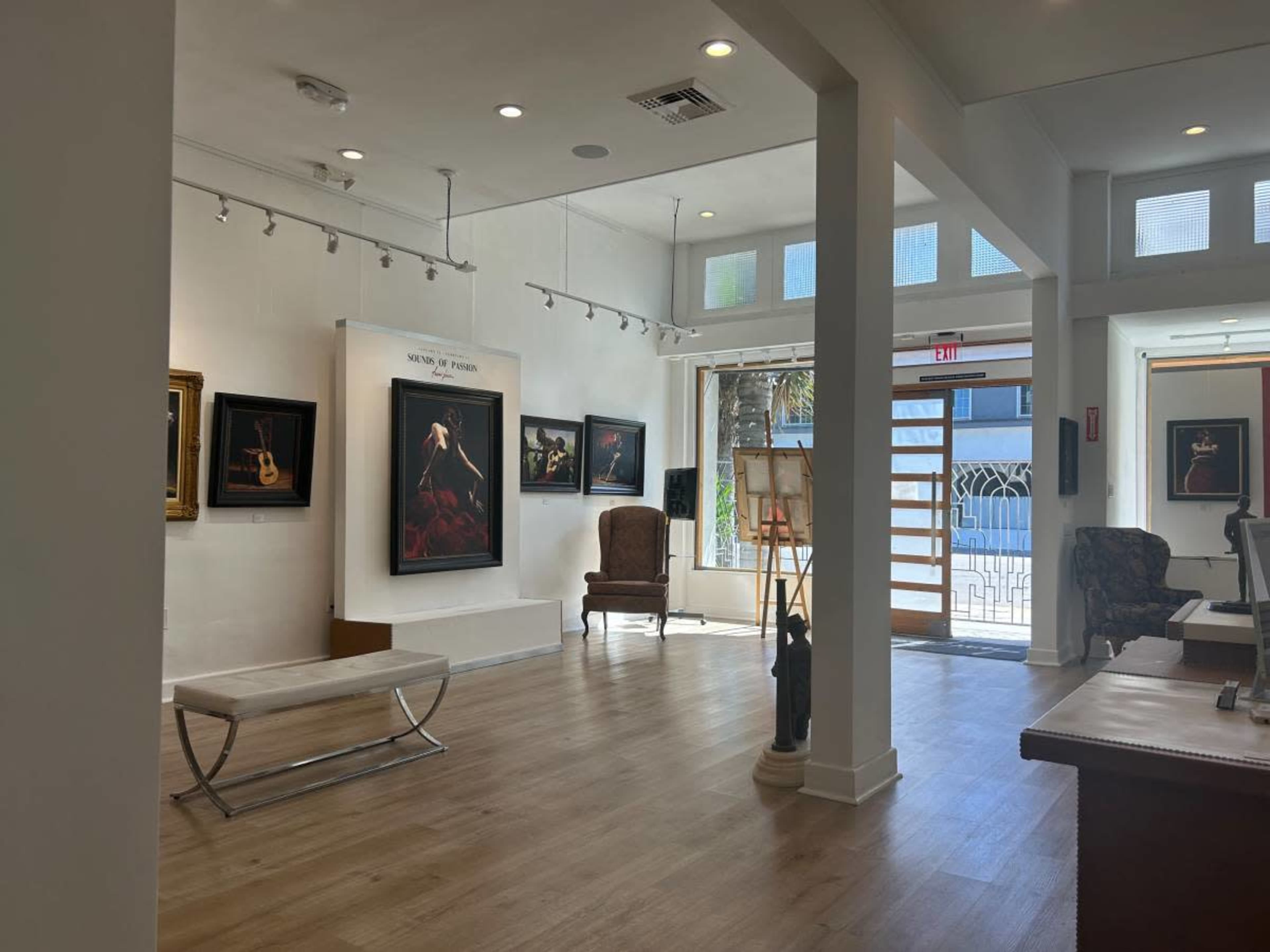The image shows a well-lit art gallery featuring framed paintings on the walls, a wooden chair, and a reception area with a bench and easel.