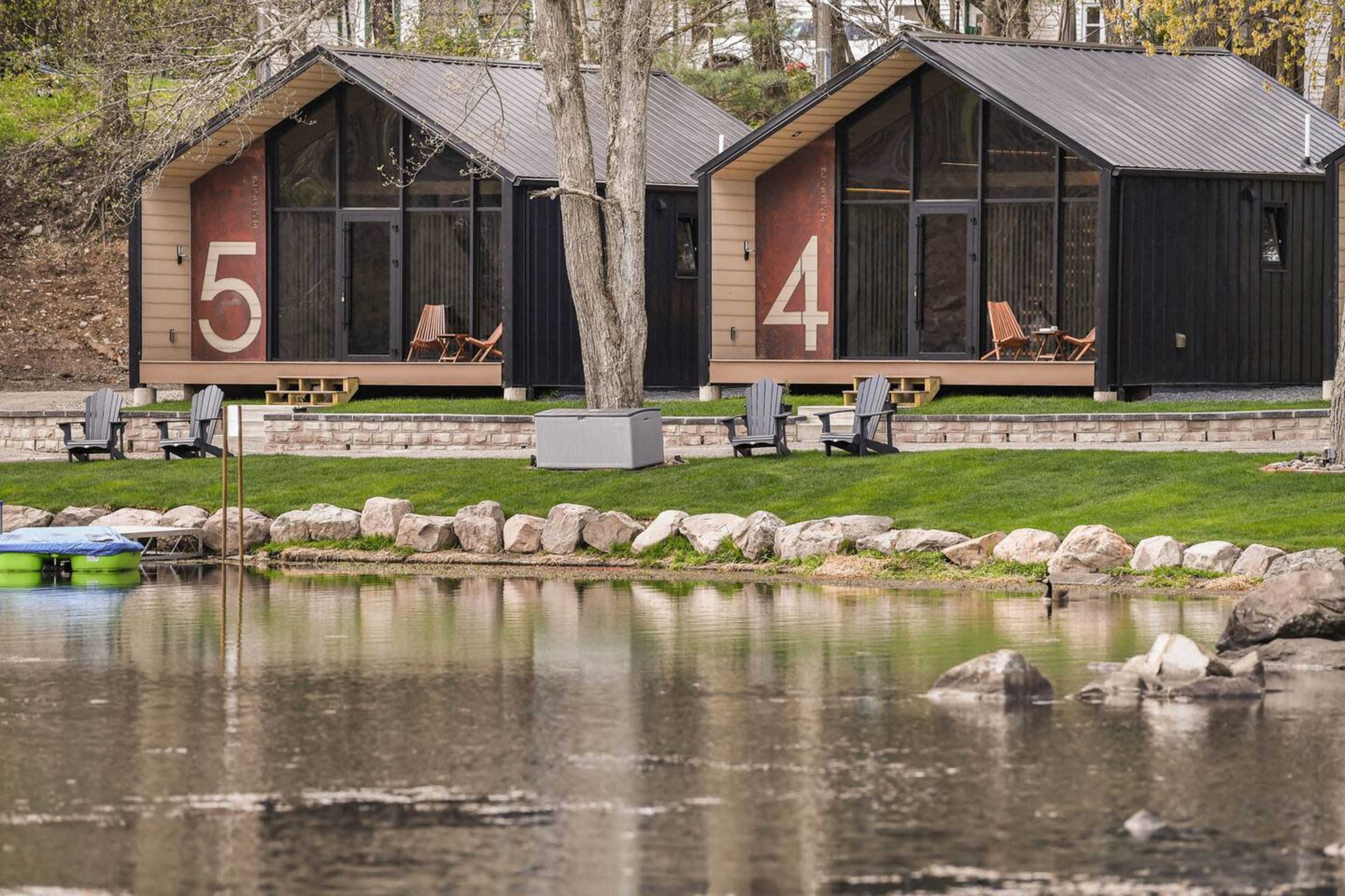 Two modern cabins numbered 4 and 5 beside a calm lake, with seating areas on their decks and a grassy lawn in front.