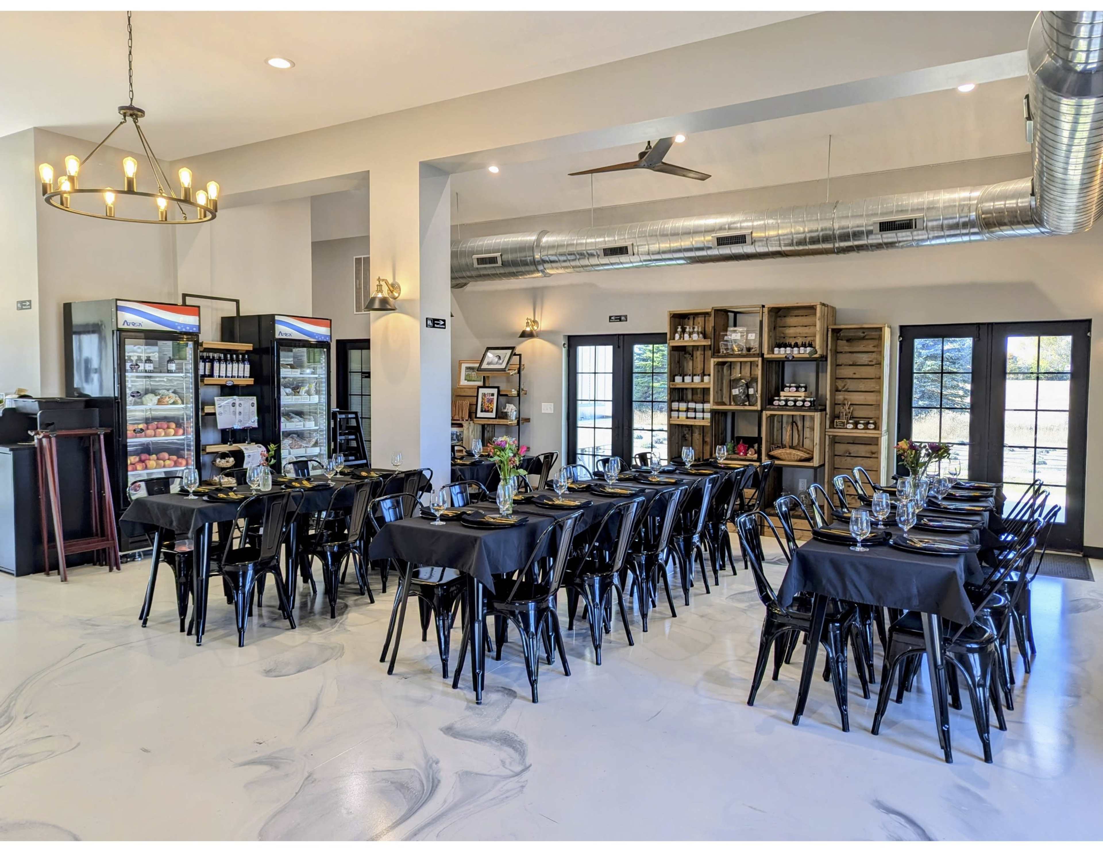 The image shows a modern dining area with black tables and chairs, industrial lighting, and a display of food and beverages in glass refrigerators.
