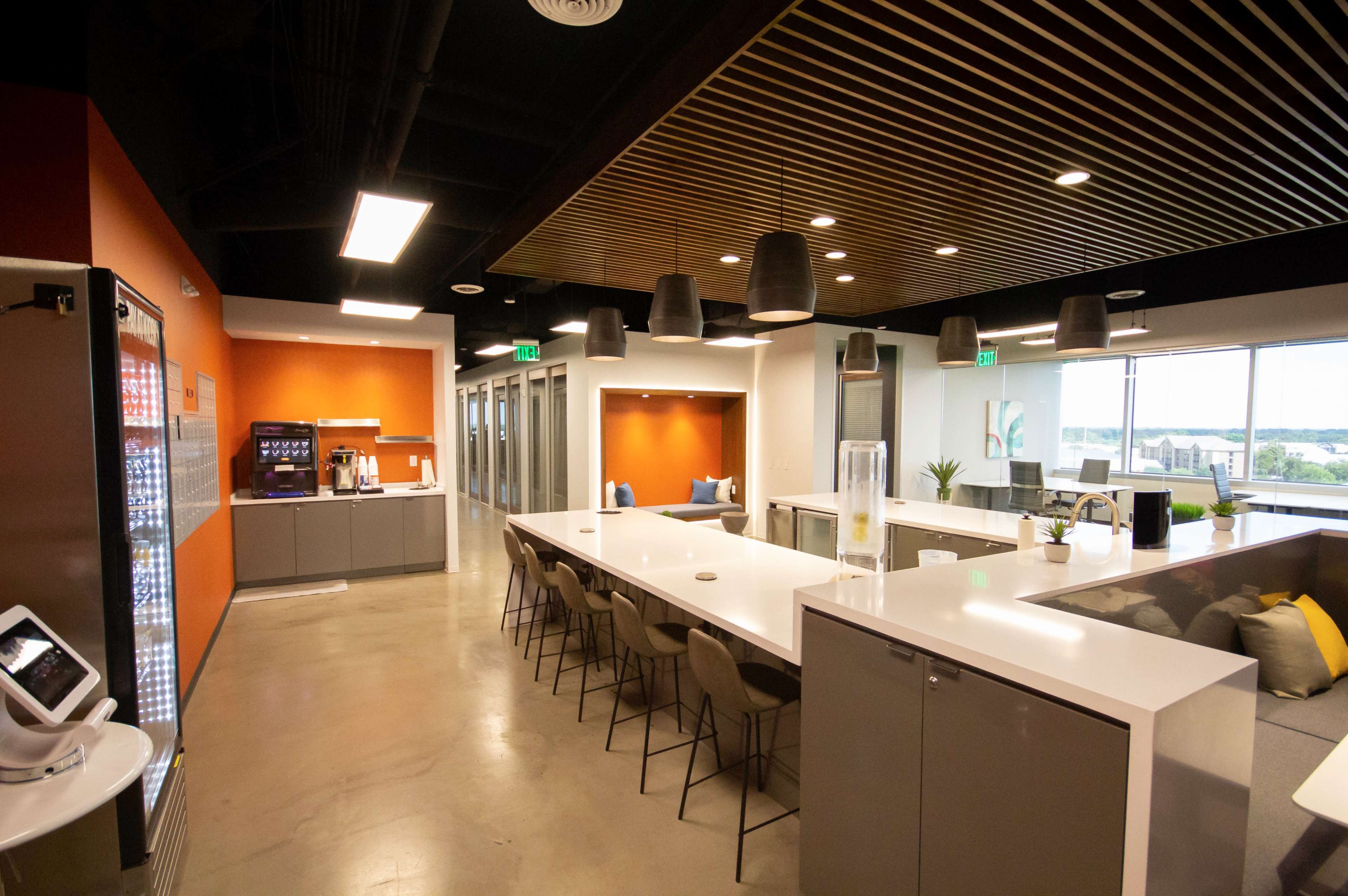A modern office kitchen and lounge area with a long white countertop, bar stools, and large windows overlooking a green landscape.