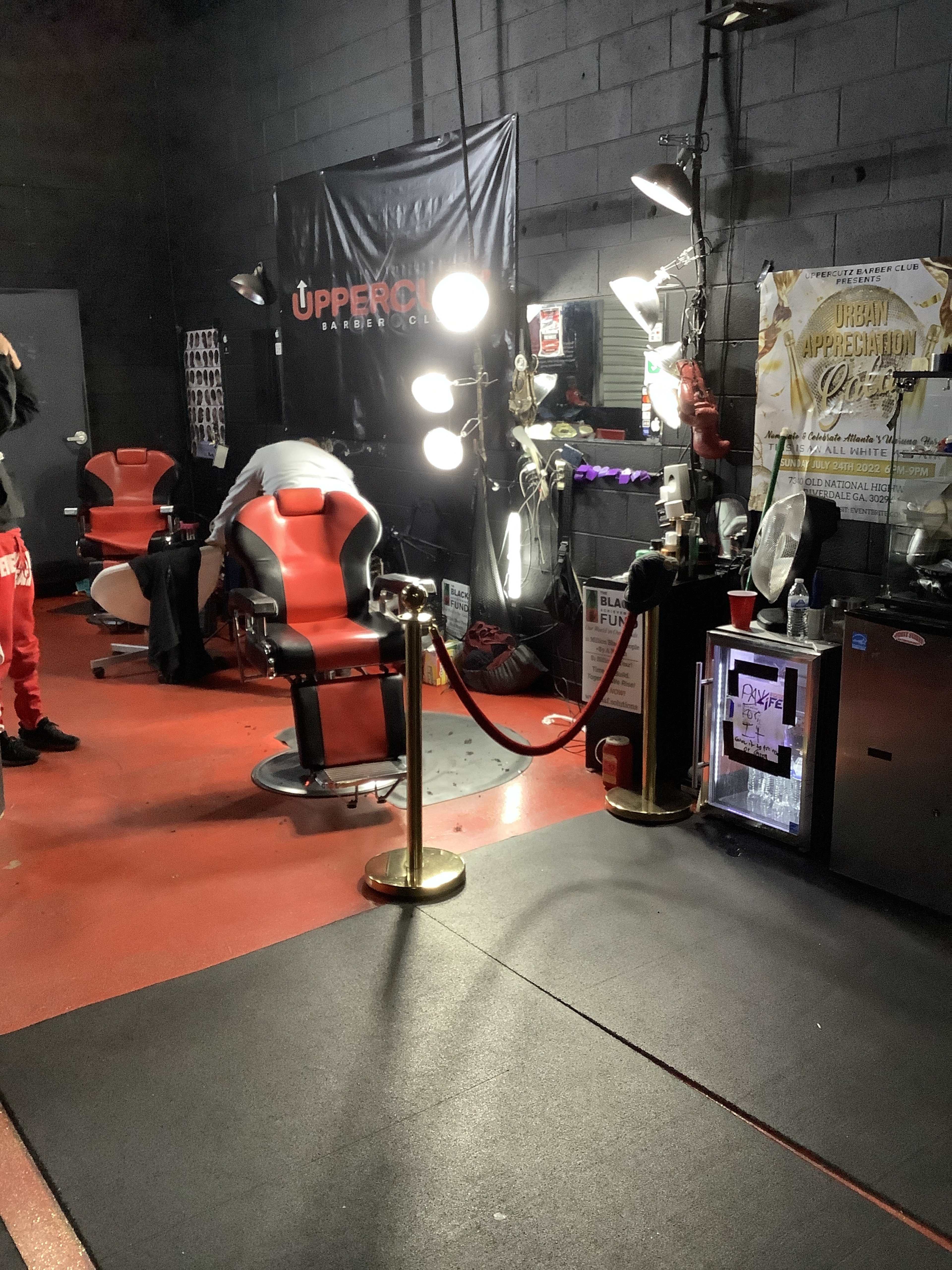 A barbershop interior with two red and black barber chairs, a waiting area with a rope barrier, and various barber tools and equipment visible on the walls.