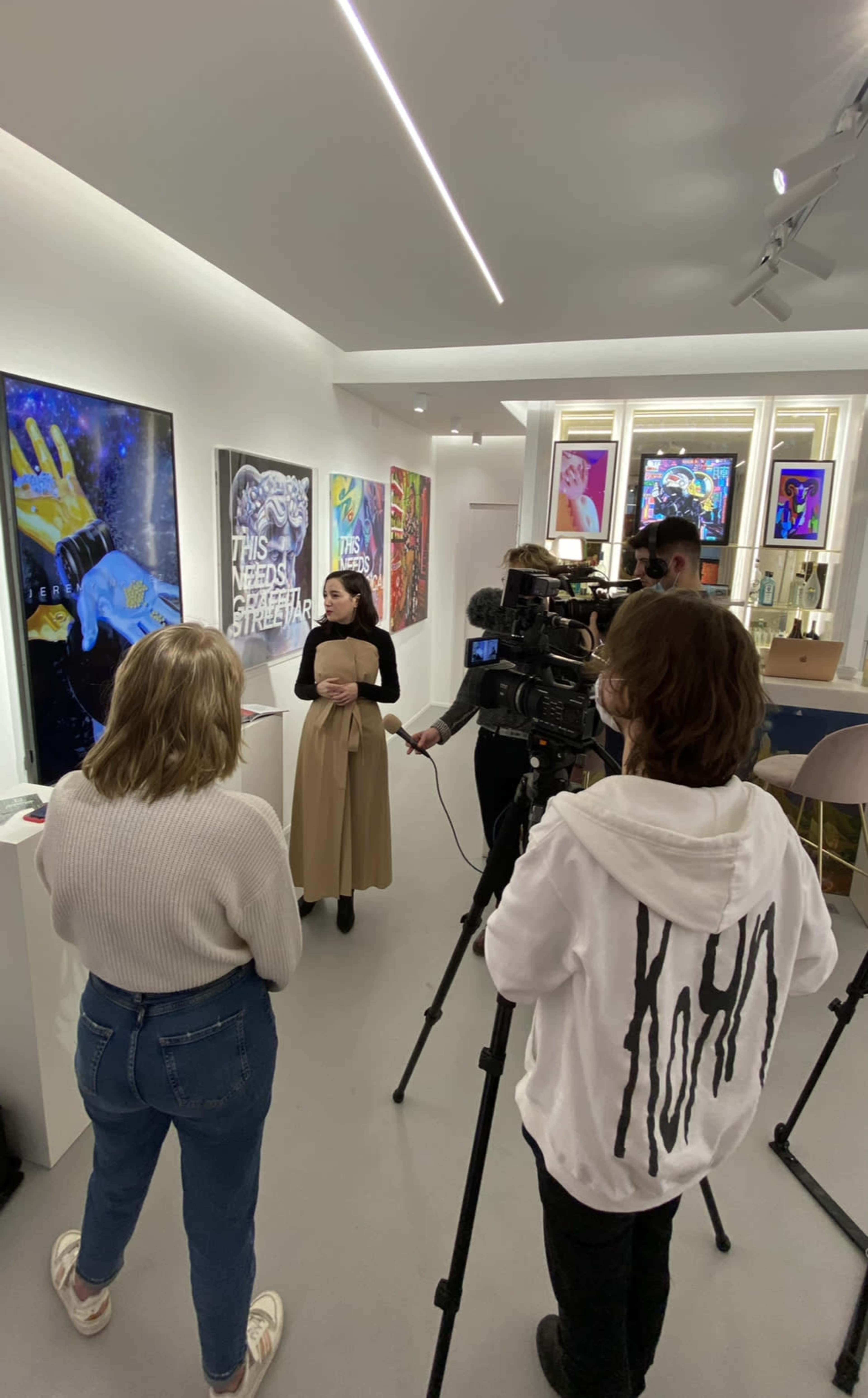 A woman is being interviewed in an art gallery with various colorful artworks displayed on the walls.
