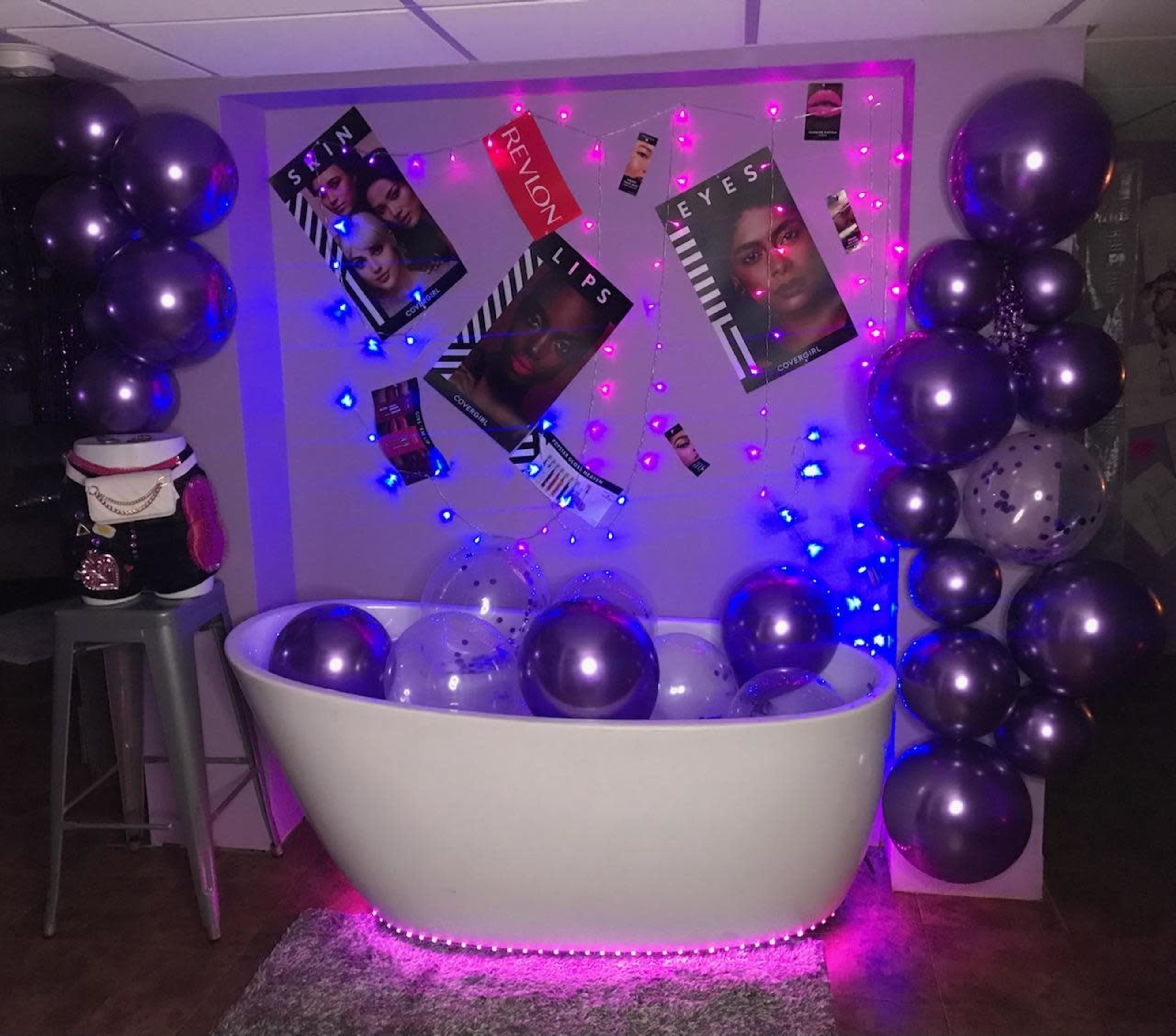 A white bathtub is surrounded by purple and blue balloons and posters promoting Revlon products, with LED lights highlighting the scene.