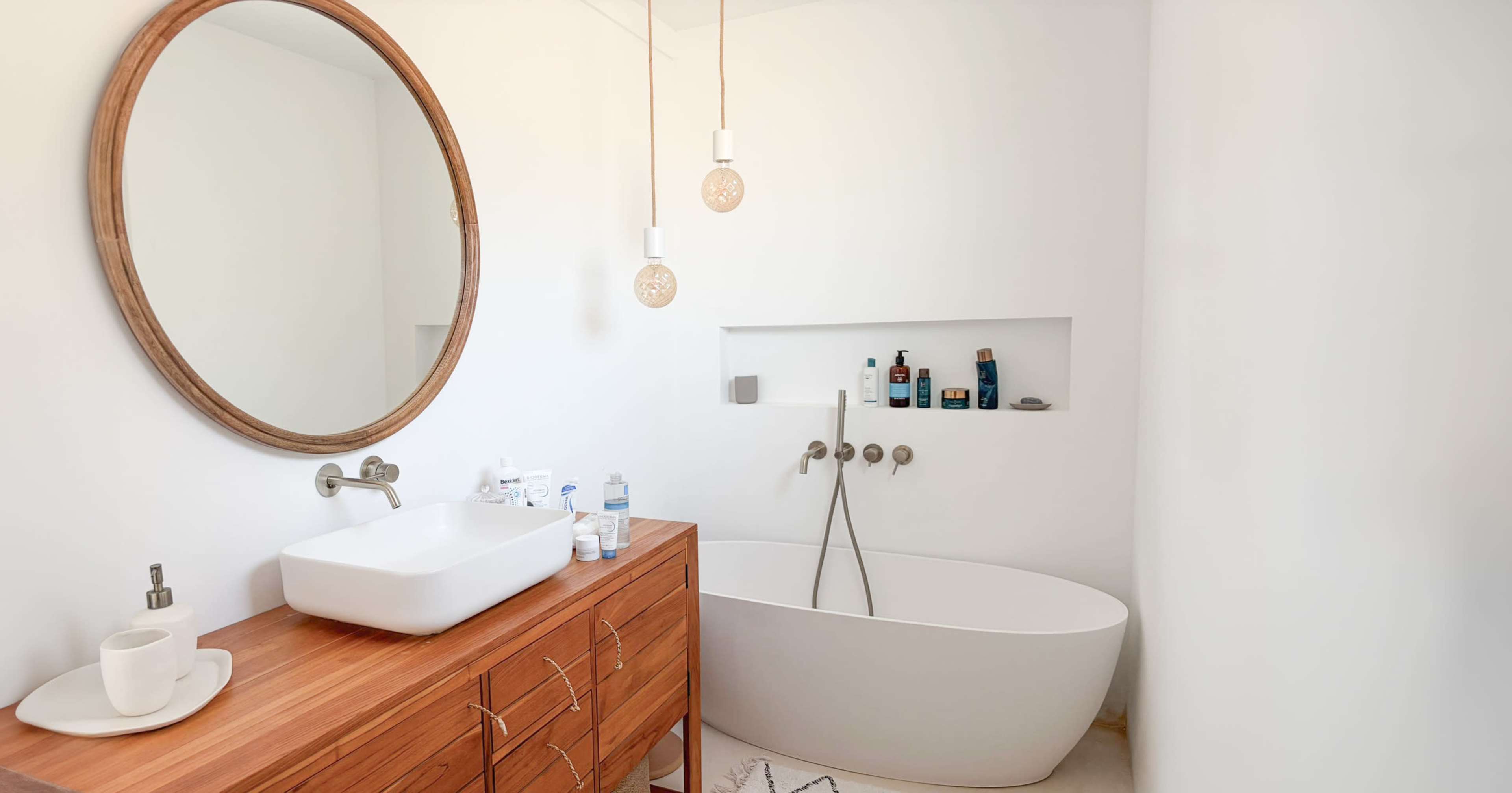 The image shows a modern bathroom featuring a round mirror, a wooden vanity with a sink, a freestanding bathtub, and wall-mounted shelves with toiletries.