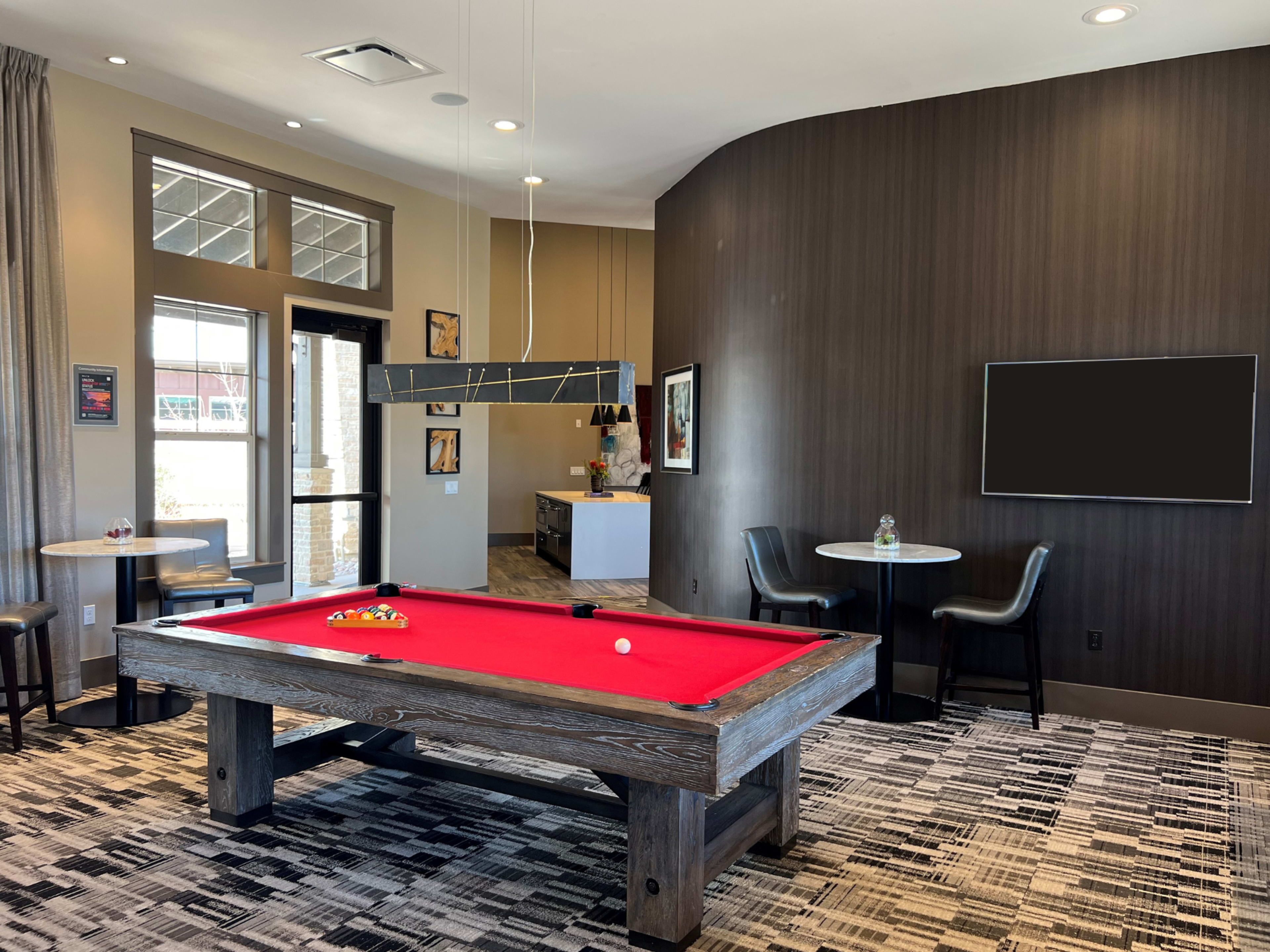 A billiards room featuring a red pool table with a wooden frame, surrounded by chairs and tables, and a wall-mounted television in the background.