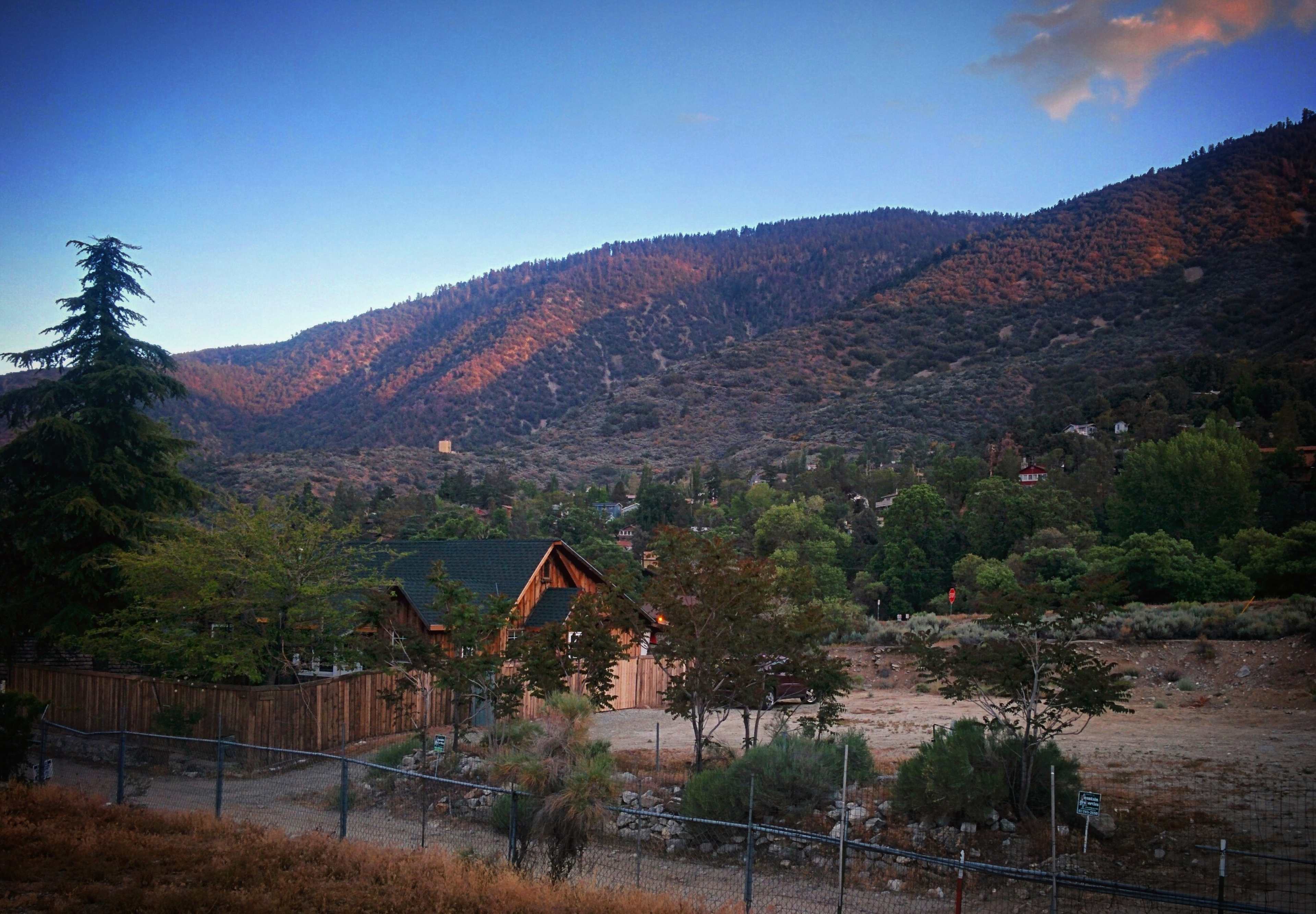 A wooden house is situated in a valley surrounded by mountains and scattered trees under a dusk sky.