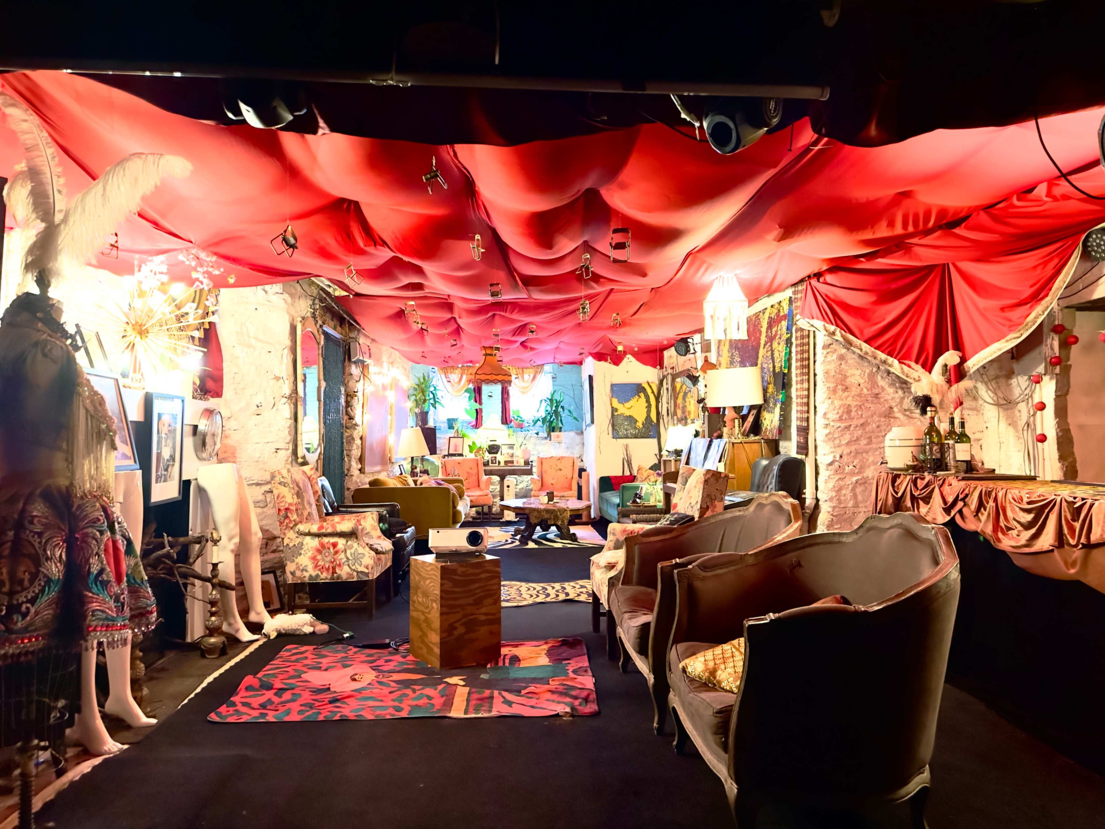 The image shows a creatively decorated interior space with red fabric overhead, various eclectic furnishings, and art adorning the walls.