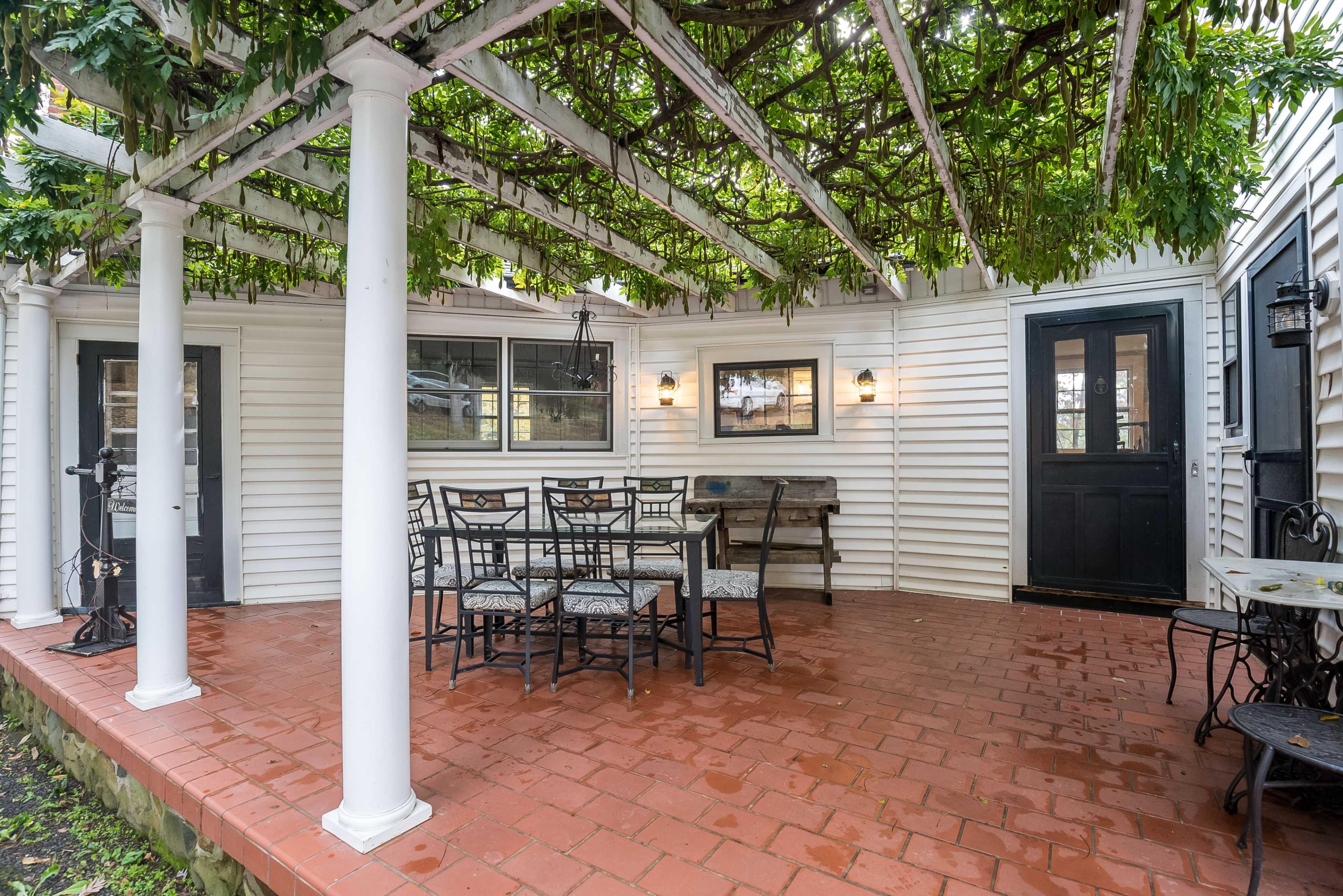 A covered patio with a wooden trellis supports lush greenery, featuring a table and chairs set on tiled flooring.