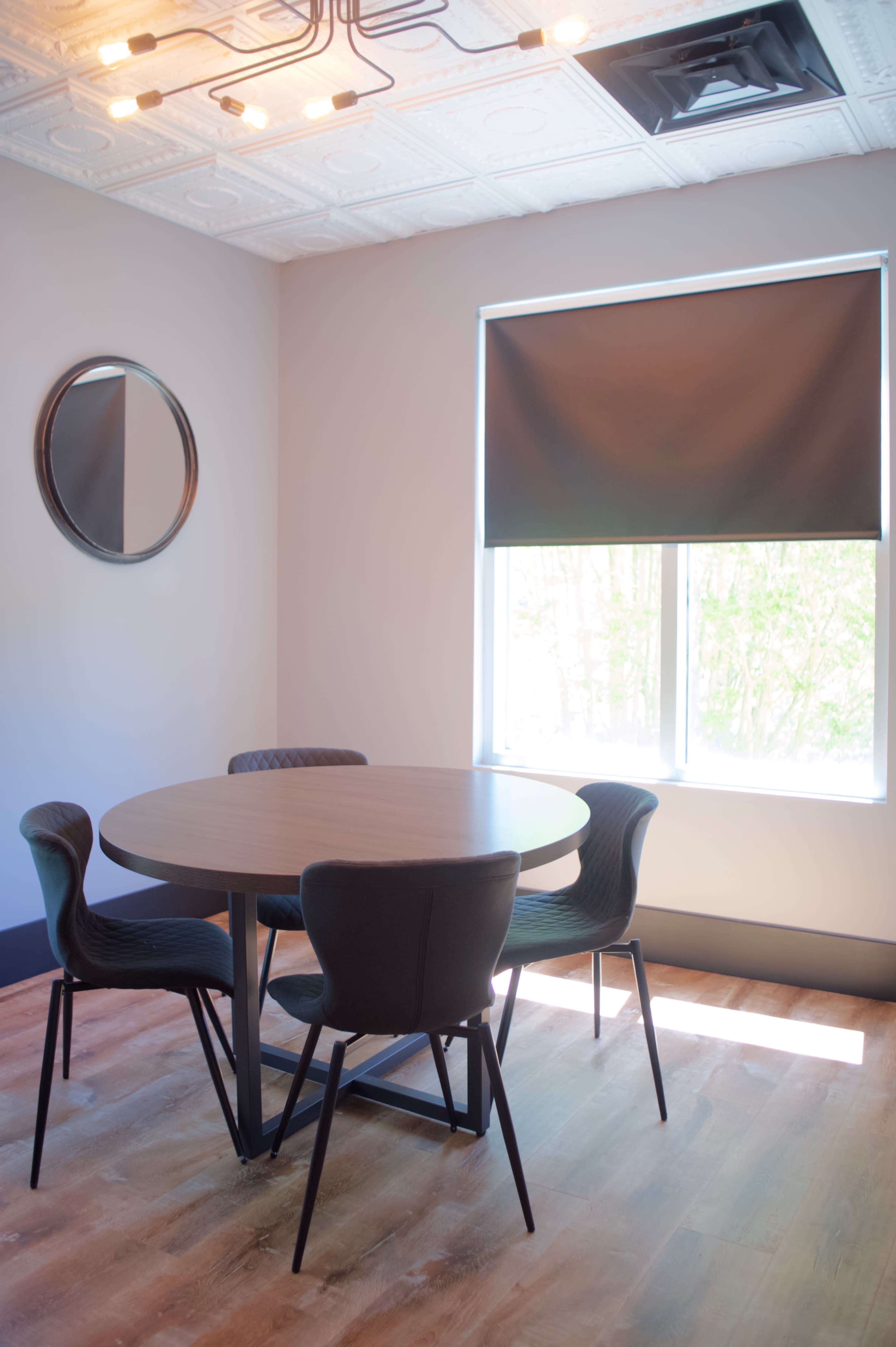 A round wooden table is surrounded by four black chairs in a room with a large window and a circular mirror on the wall.