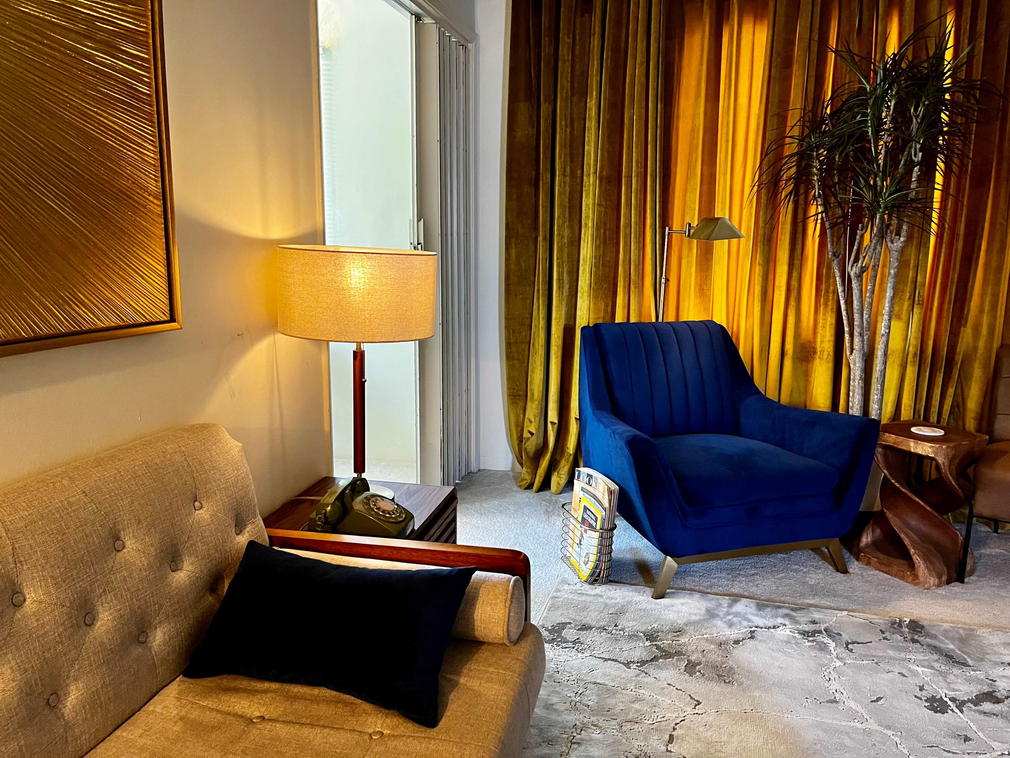 A modern living room features a beige sofa, a blue armchair, a floor lamp, and a large golden curtain in the background.