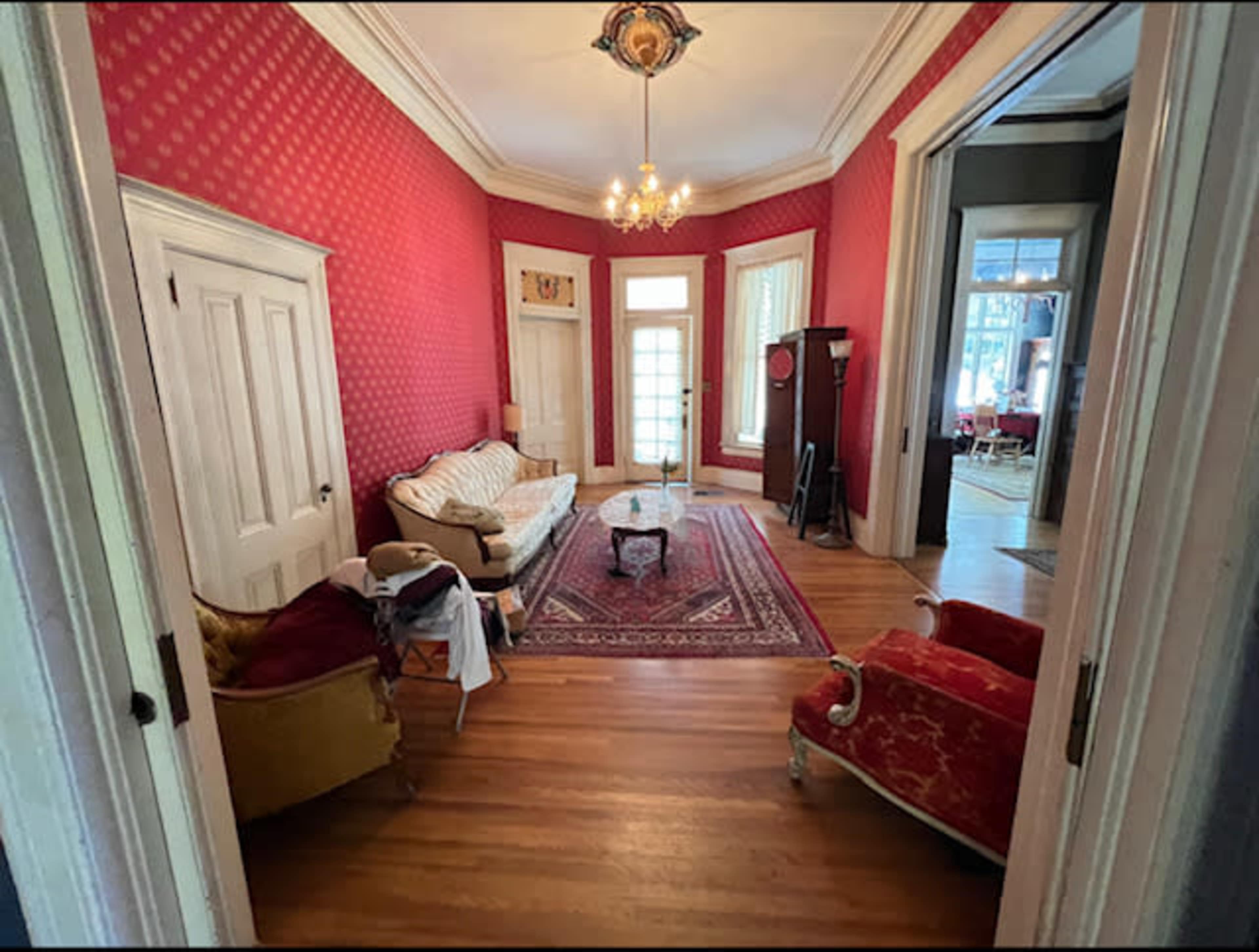 A room with red patterned walls, a wooden floor, and a vintage seating arrangement including a sofa and two chairs, centered around a small table on a patterned rug.