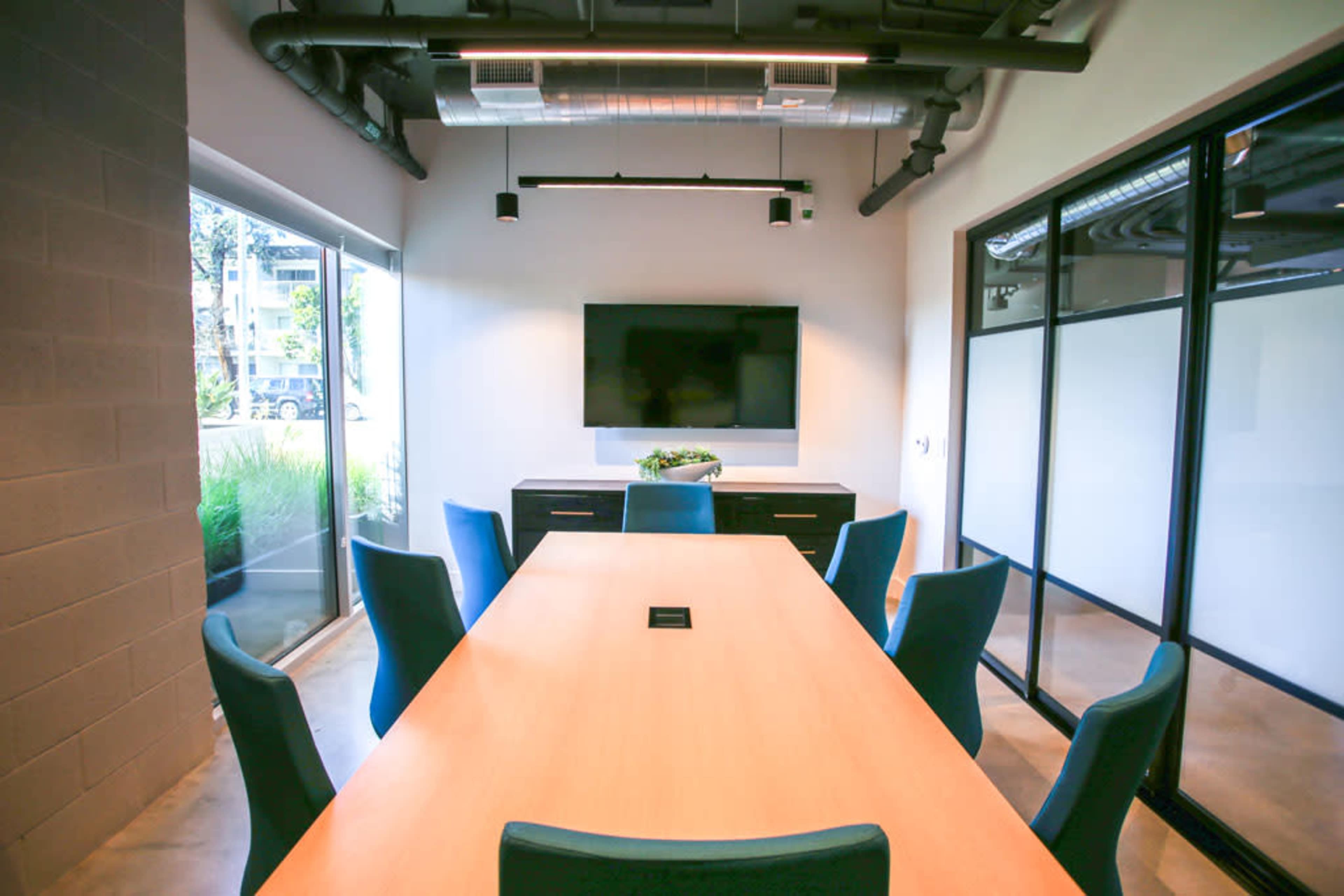 A conference room features a long wooden table surrounded by blue chairs, with a television mounted on the wall and large windows allowing natural light.