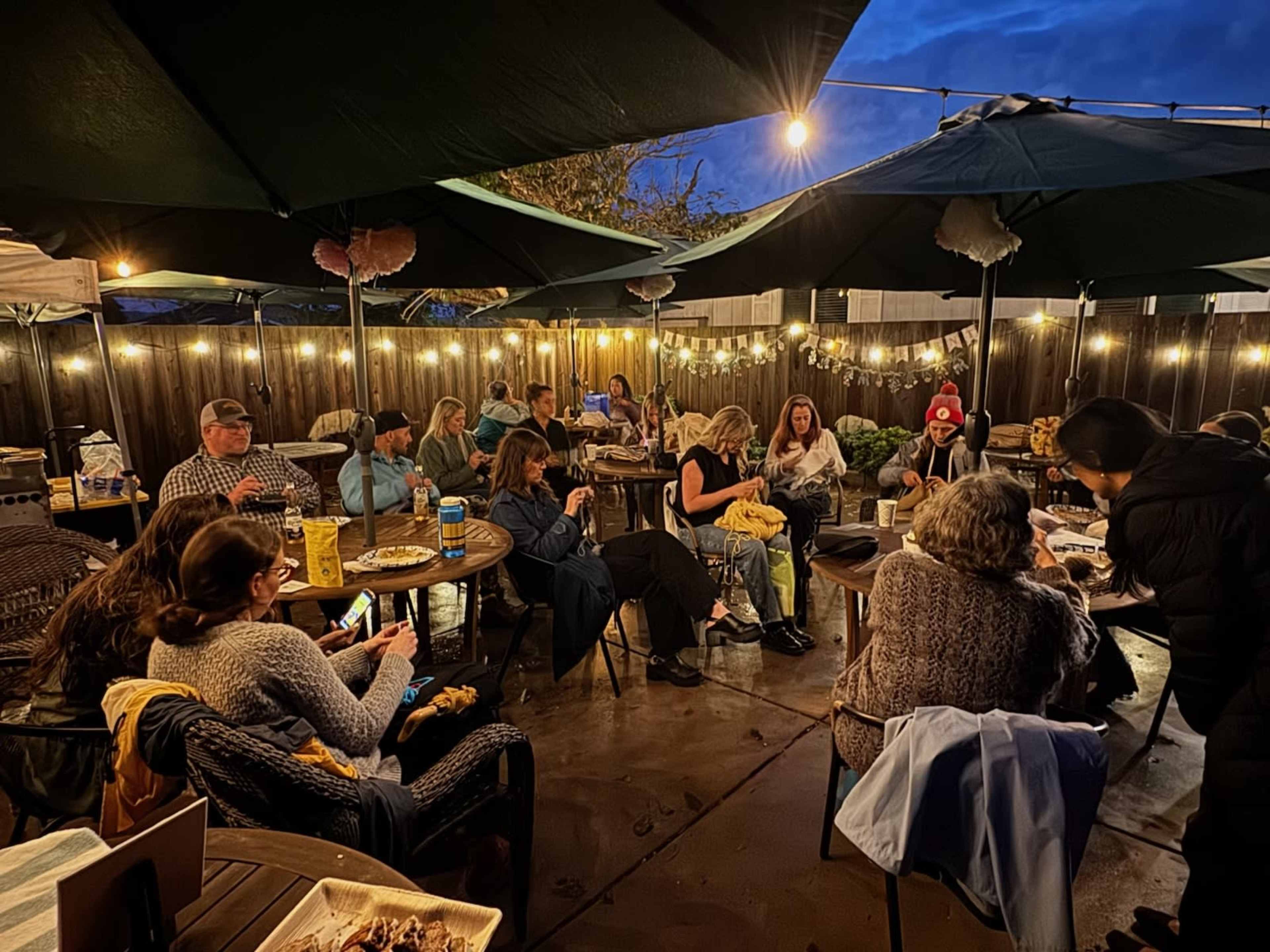 A group of people is gathered around tables under large umbrellas in an outdoor space, with string lights illuminating the area.