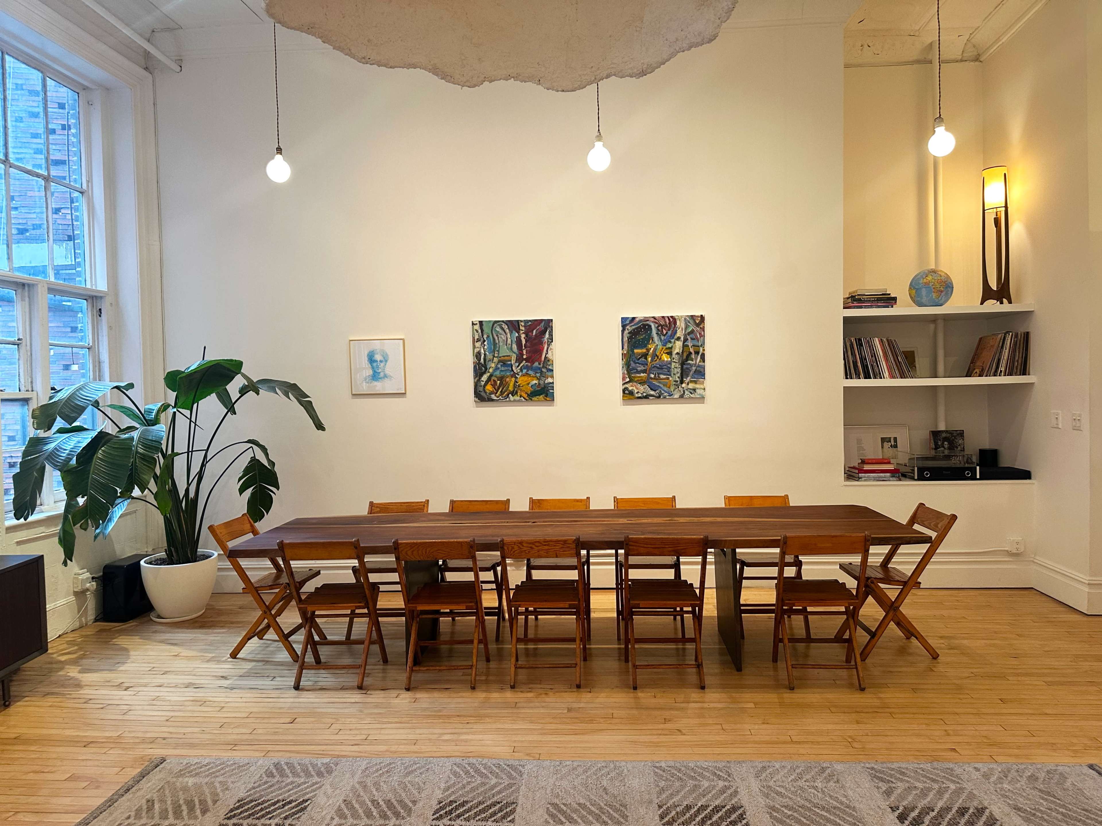 The image shows a spacious room with a large wooden dining table surrounded by folding chairs, two artworks on the wall, a tall plant in a pot, and shelves with books and a globe.