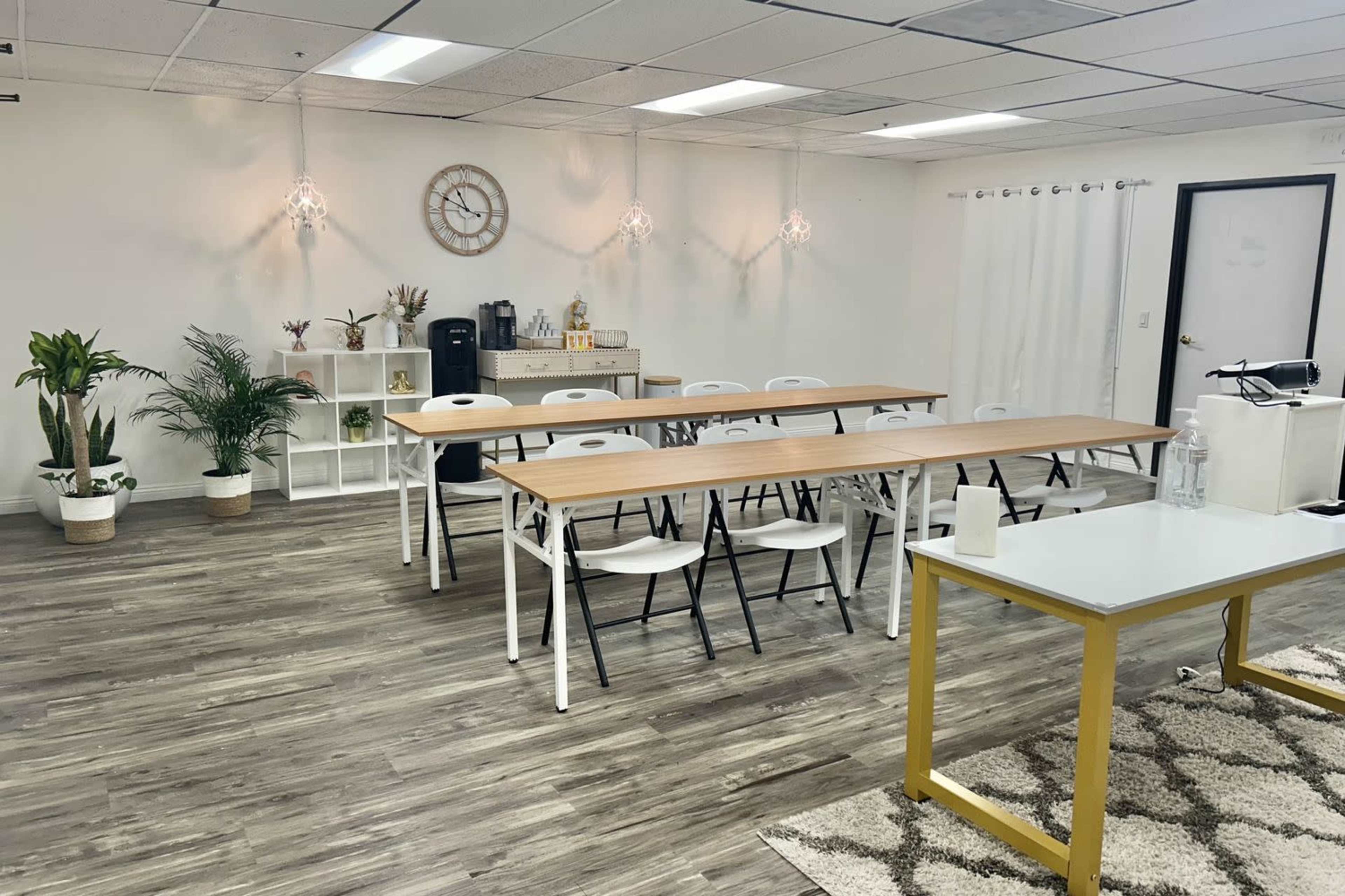 The image shows a modern meeting room featuring several wooden tables arranged with white chairs, plants, and decorative elements in a well-lit environment.