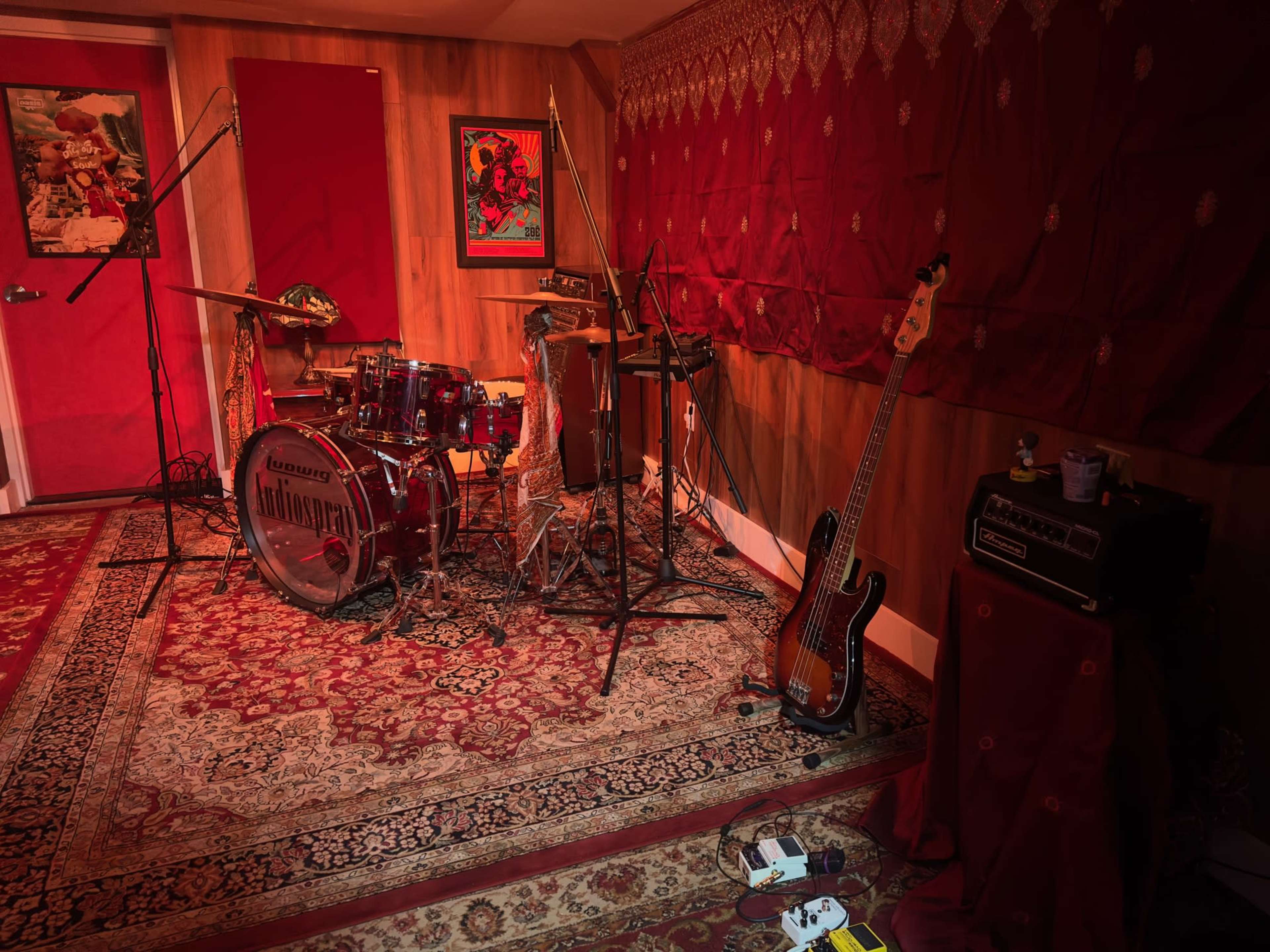 A music recording studio with a drum set, bass guitar, and various microphones arranged on red-draped walls.