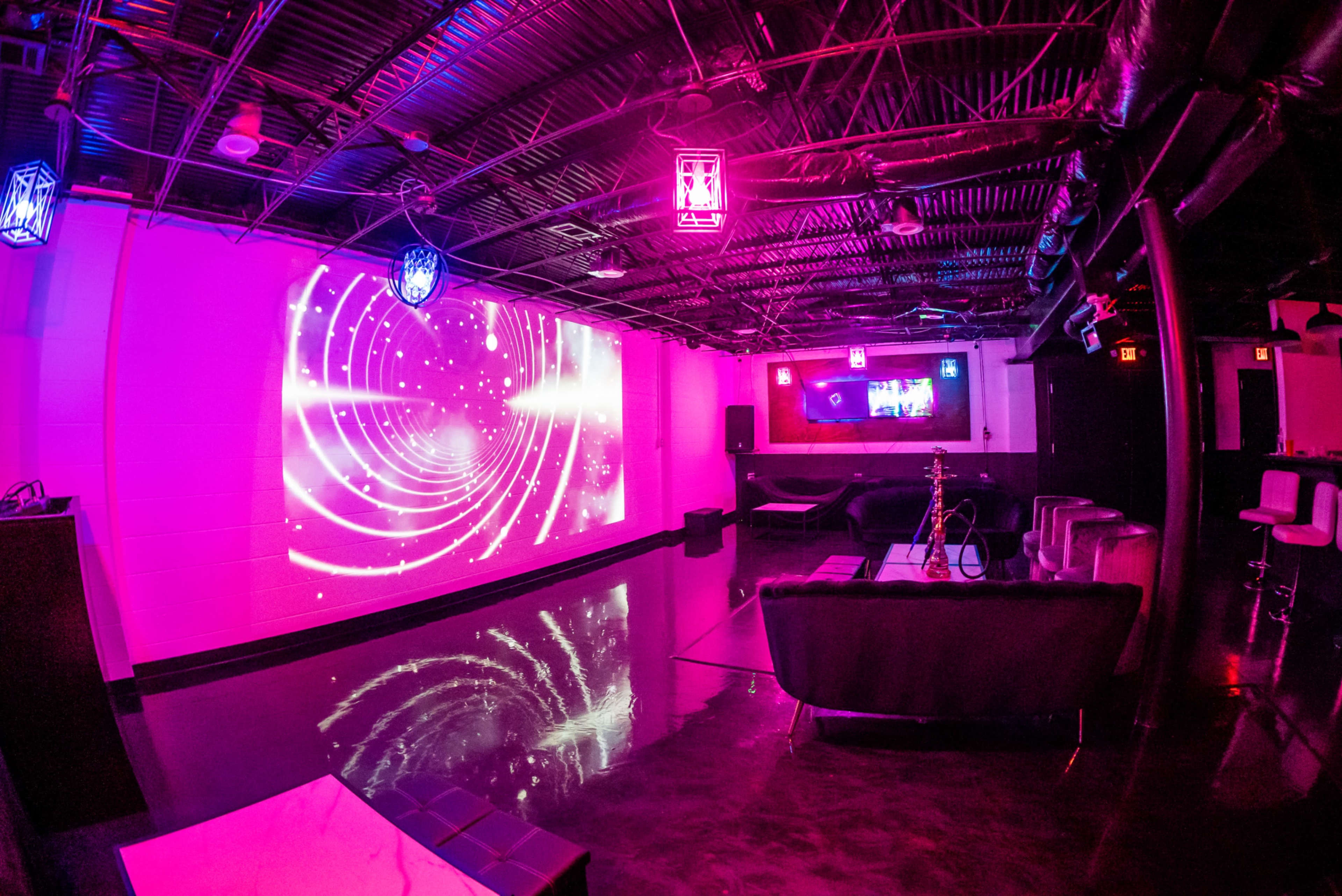 A modern lounge space with a shiny floor, illuminated by colorful lights and featuring a large projection on the wall.