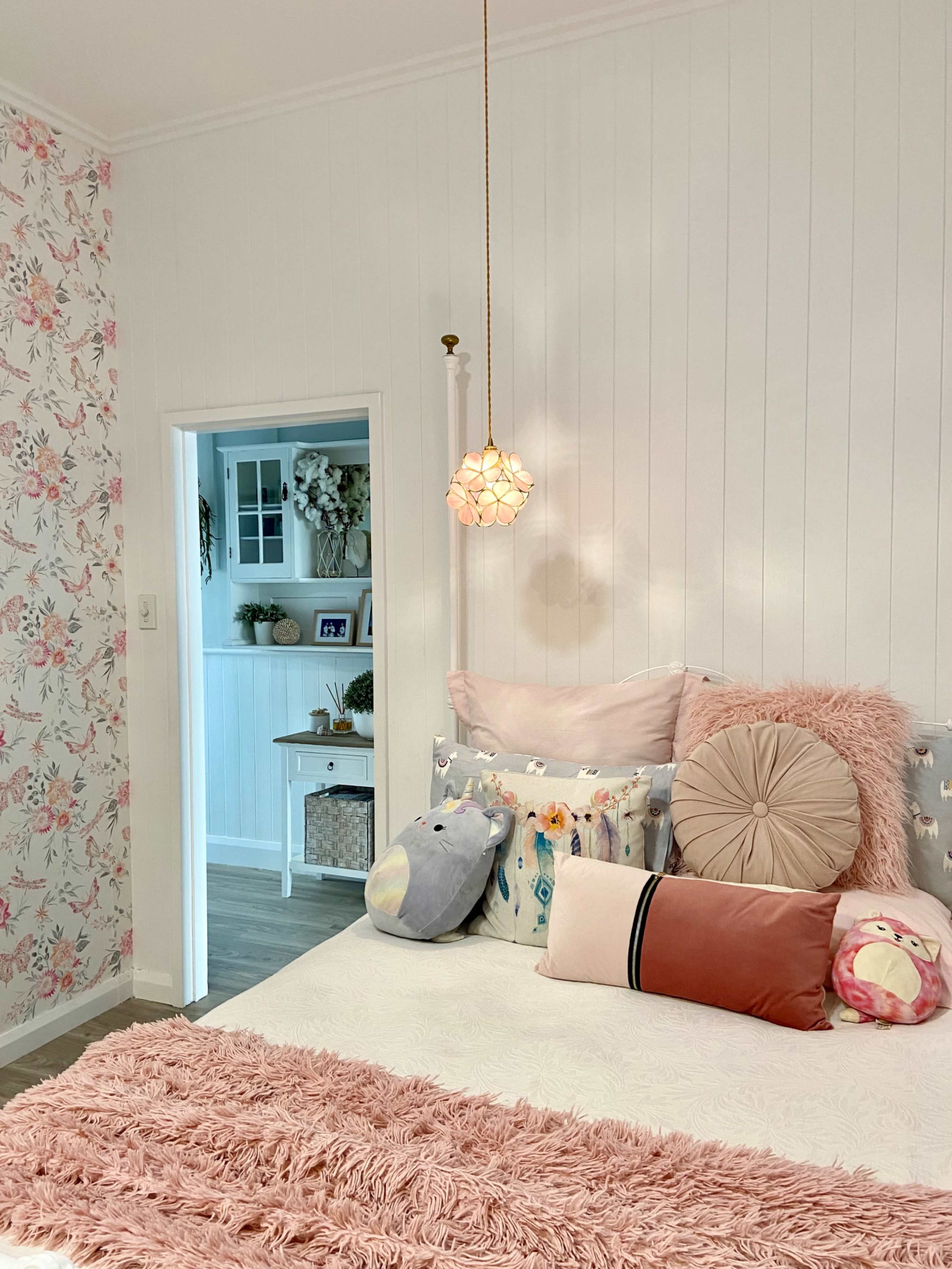 A cozy bedroom features a bed with decorative pillows and a pendant light, set against floral wallpaper and an open doorway leading to another room.