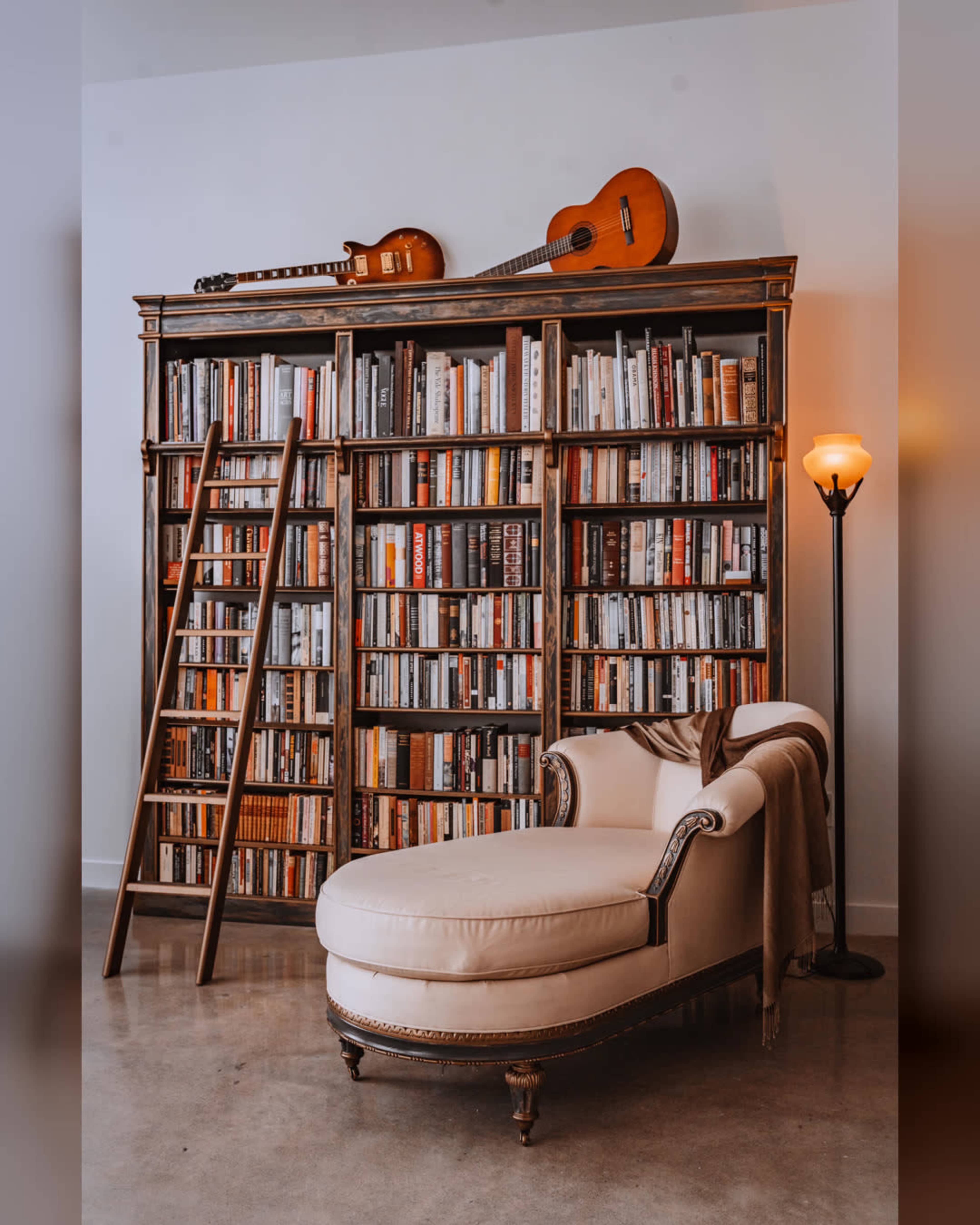 A tall bookshelf filled with books stands against the wall, accompanied by a cozy chaise lounge and two guitars positioned on top of the shelf.