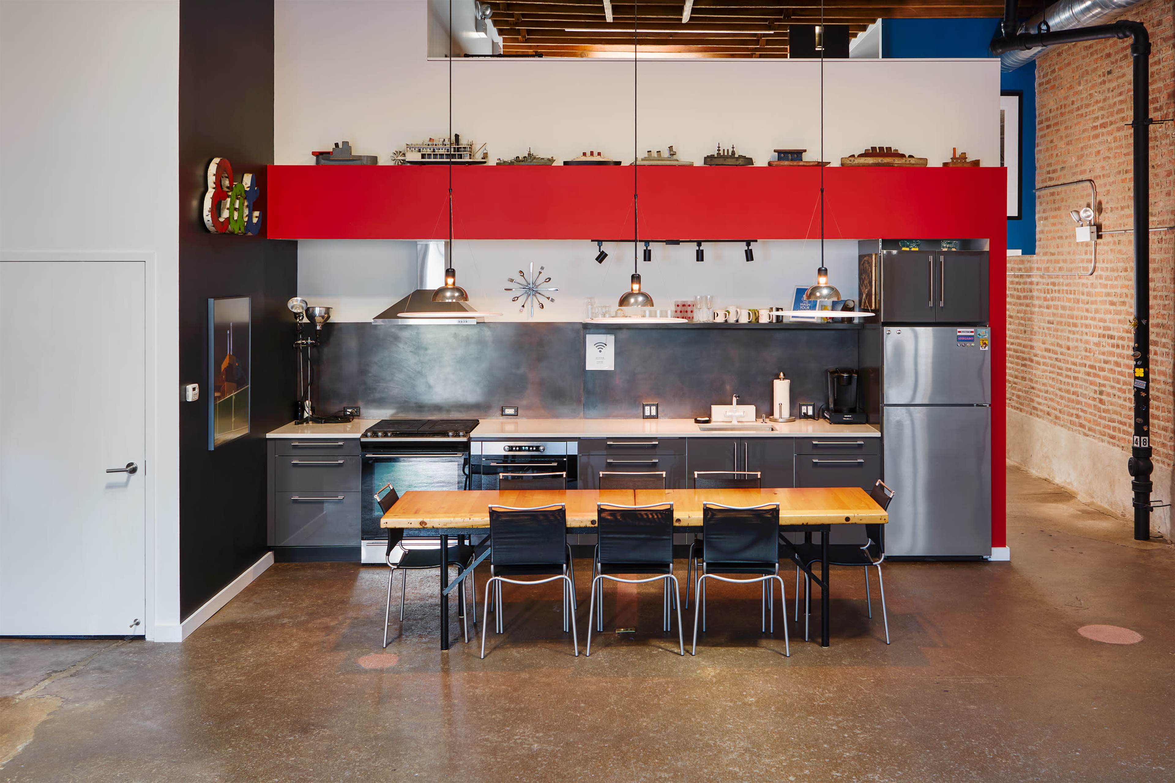 A modern kitchen with a long wooden table and black chairs, featuring red and gray cabinetry, industrial lighting, and stainless steel appliances.