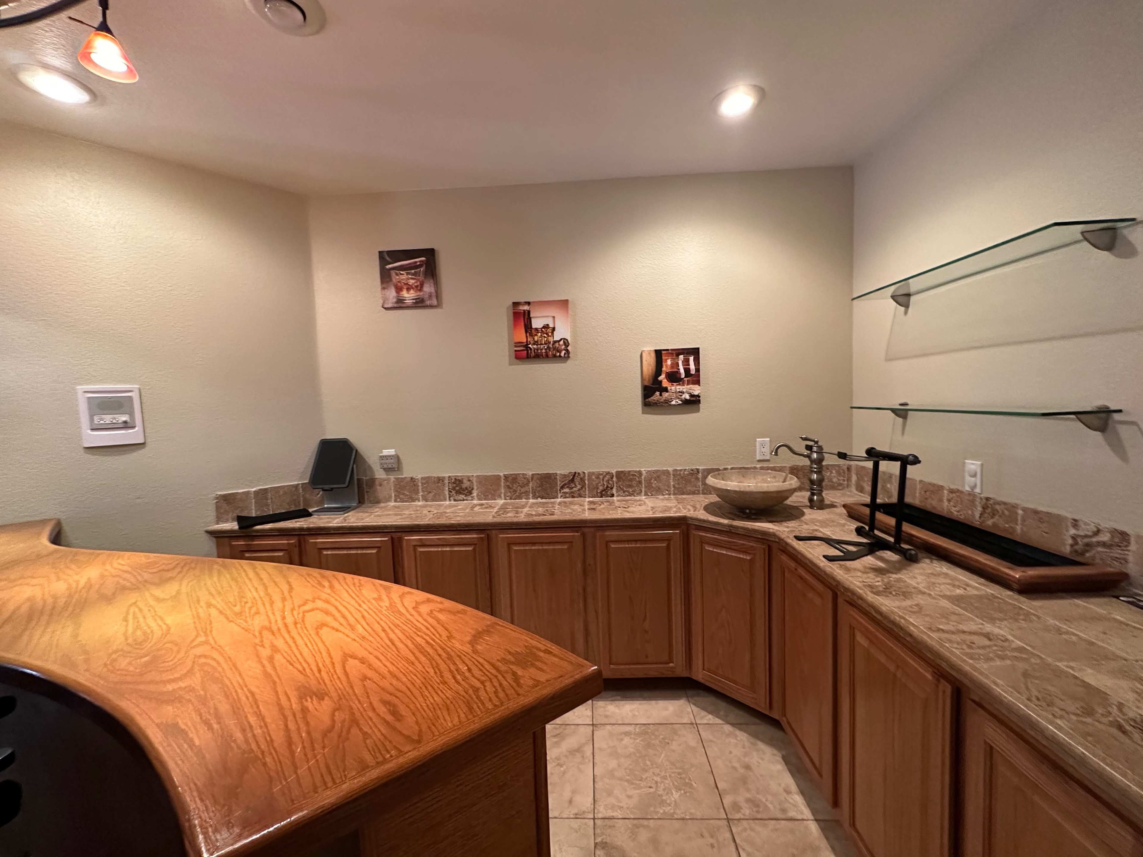 A kitchen area with wooden cabinets, a tiled countertop, and wall-mounted shelves displaying framed pictures.