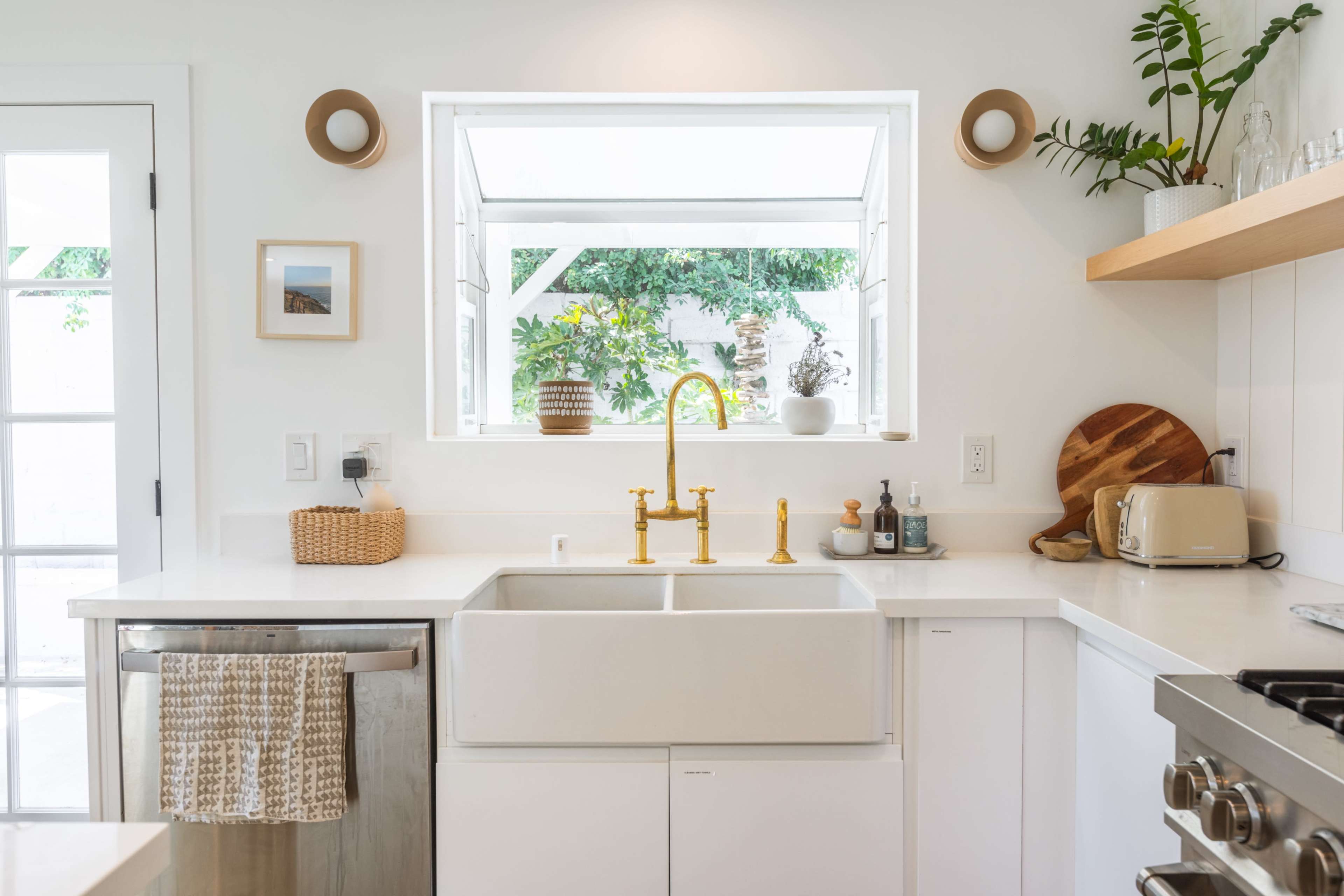 A modern kitchen features a central farmhouse sink with a gold faucet, surrounded by white cabinets and a view of greenery through an adjacent window.