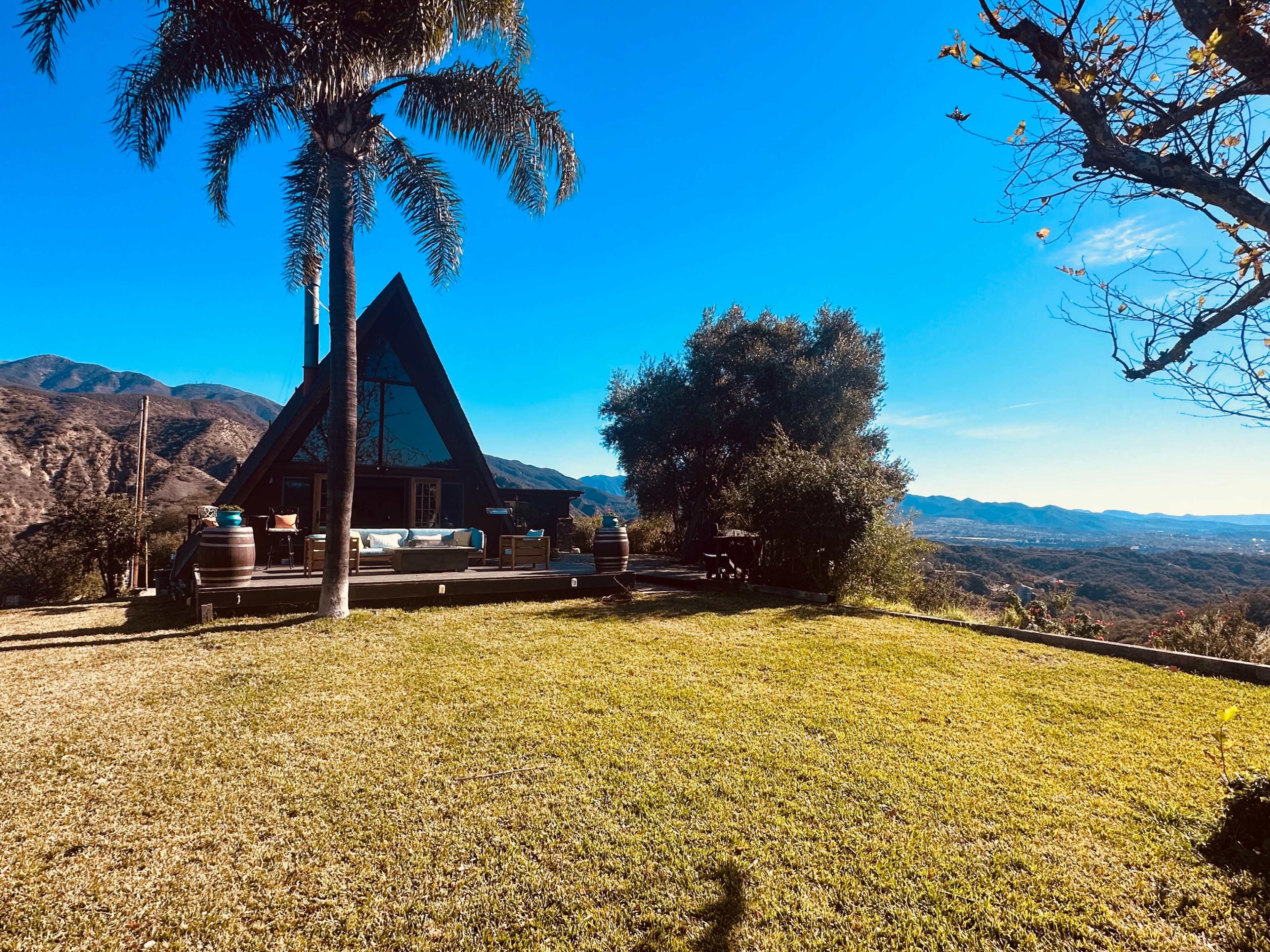A triangular-shaped cabin is situated on a grassy area overlooking a mountainous landscape under a clear blue sky.