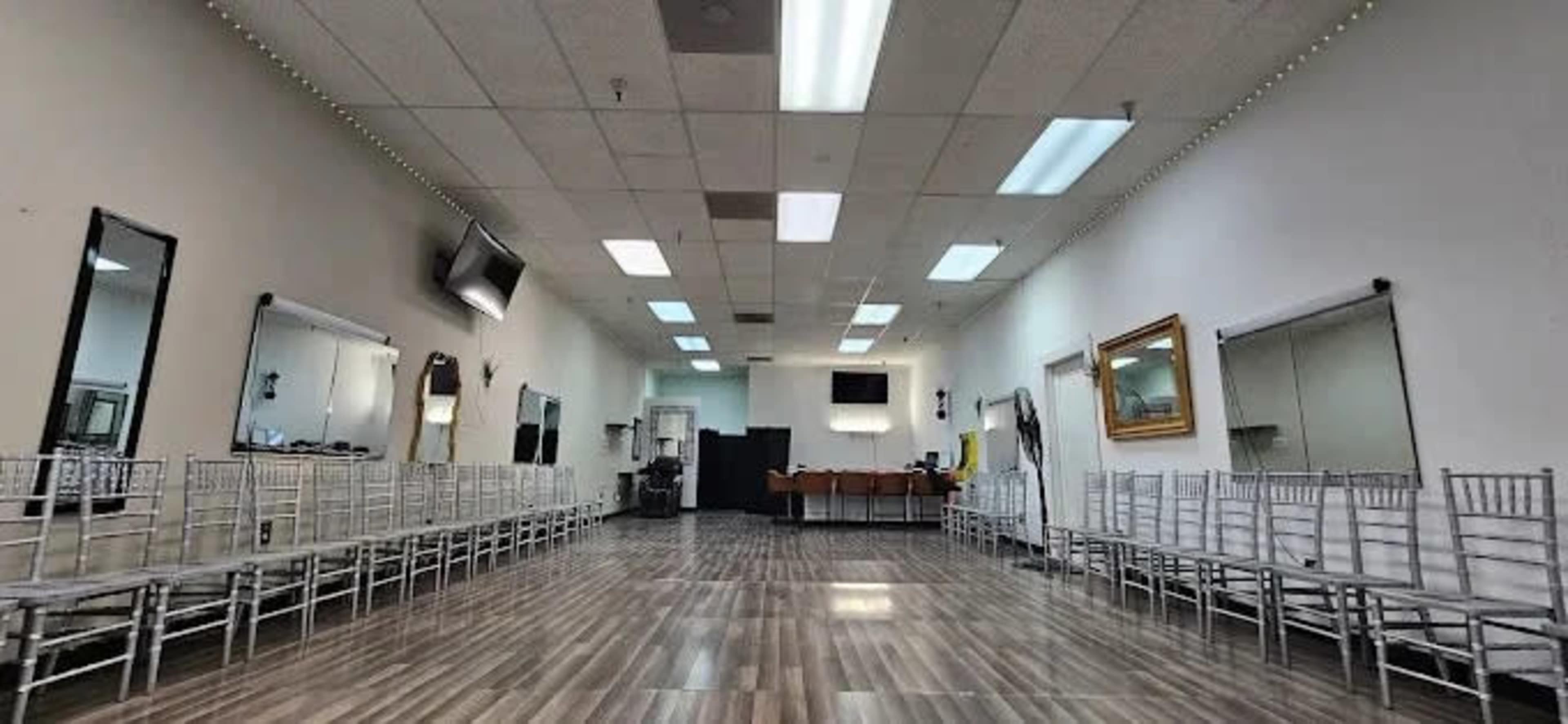 The image shows a spacious room with rows of silver chiavari chairs arranged on polished wooden flooring, along with several mirrors and a reception area at the far end.