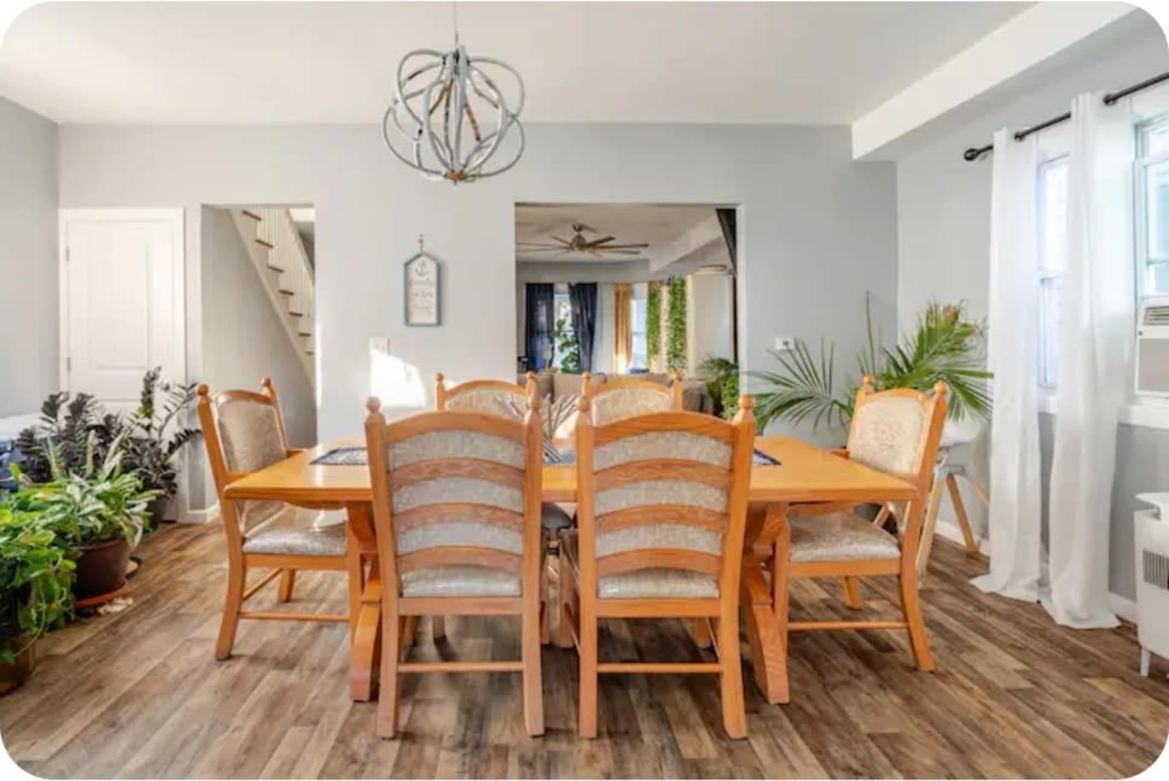 A dining room features a large wooden table surrounded by eight chairs, with windows allowing natural light and decorative plants in the background.