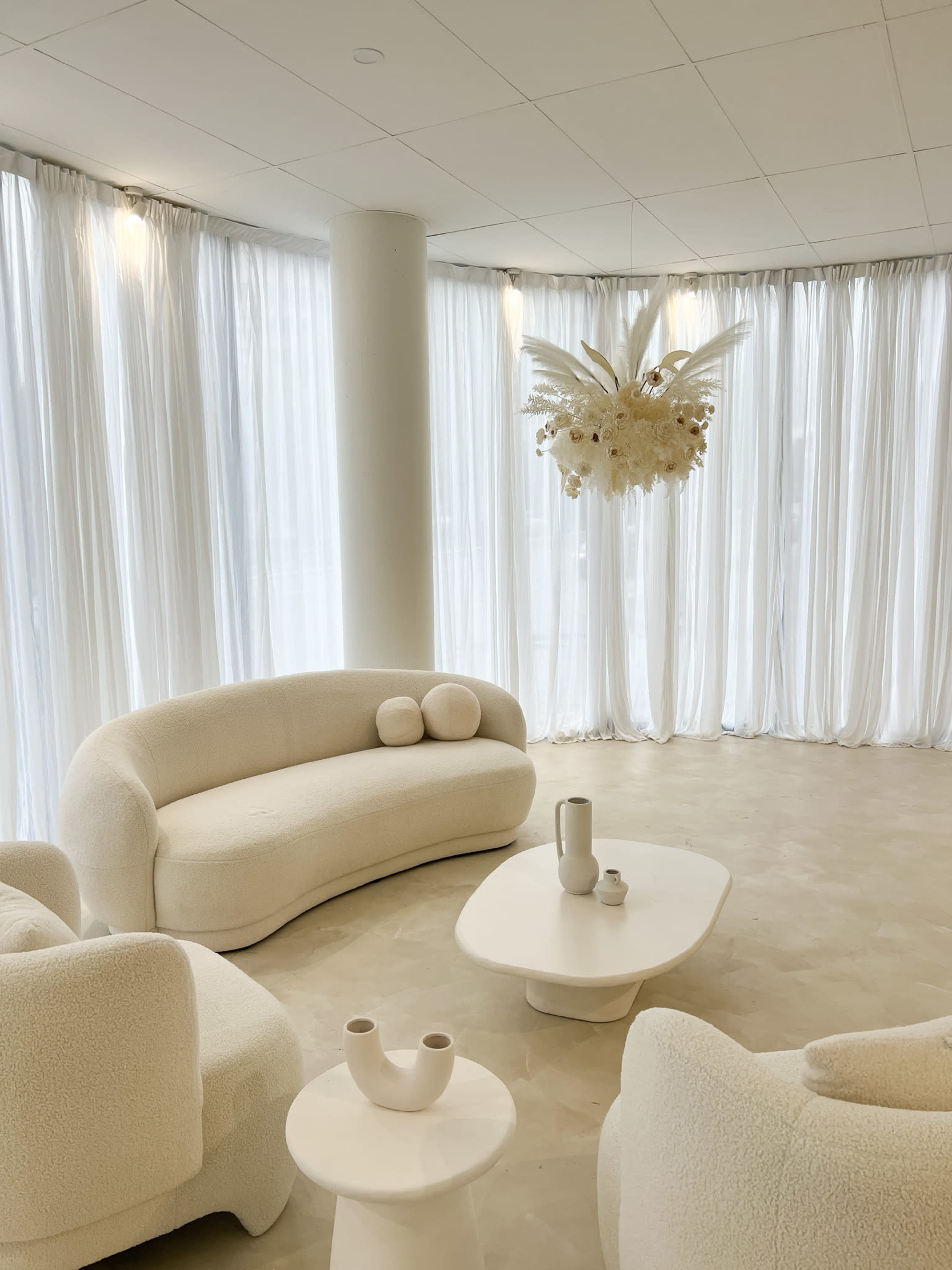 A modern, minimalist living space features white, curved furniture and sheer curtains, with a decorative light fixture overhead.