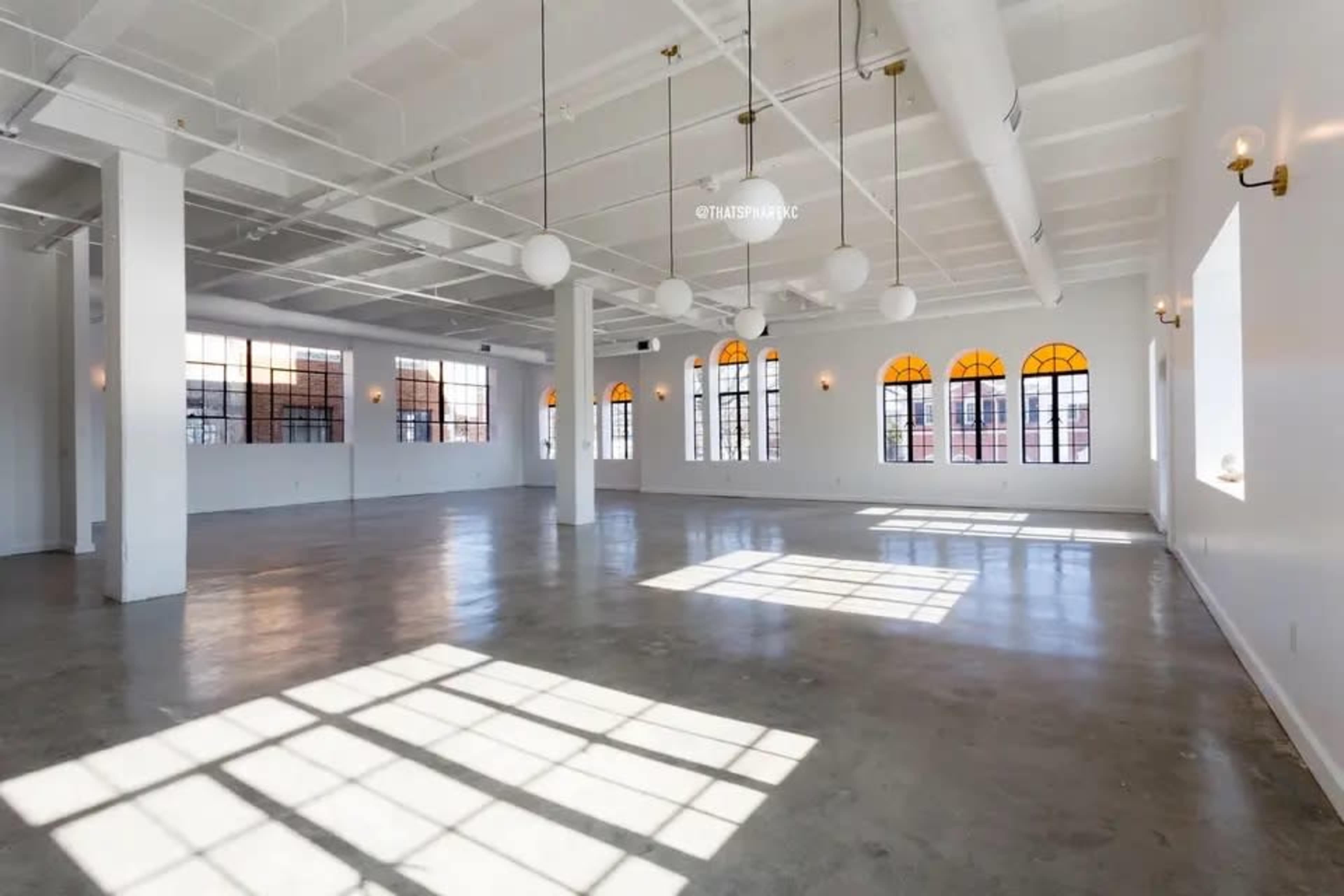 The image shows a spacious, empty room with large windows and bright, natural light casting shadows on the floor.