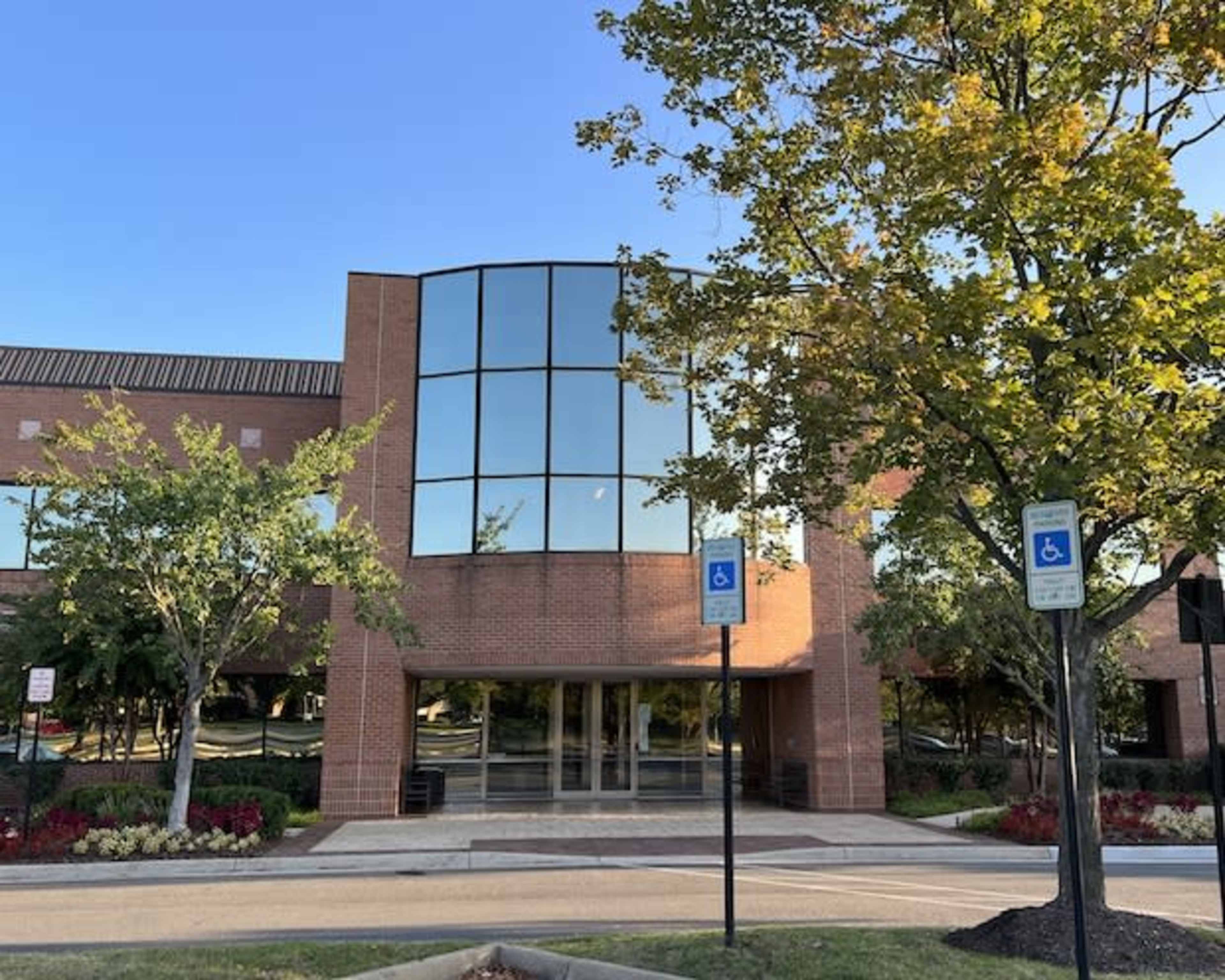 A modern brick building with large glass windows is surrounded by landscaped greenery and accessible parking spaces.
