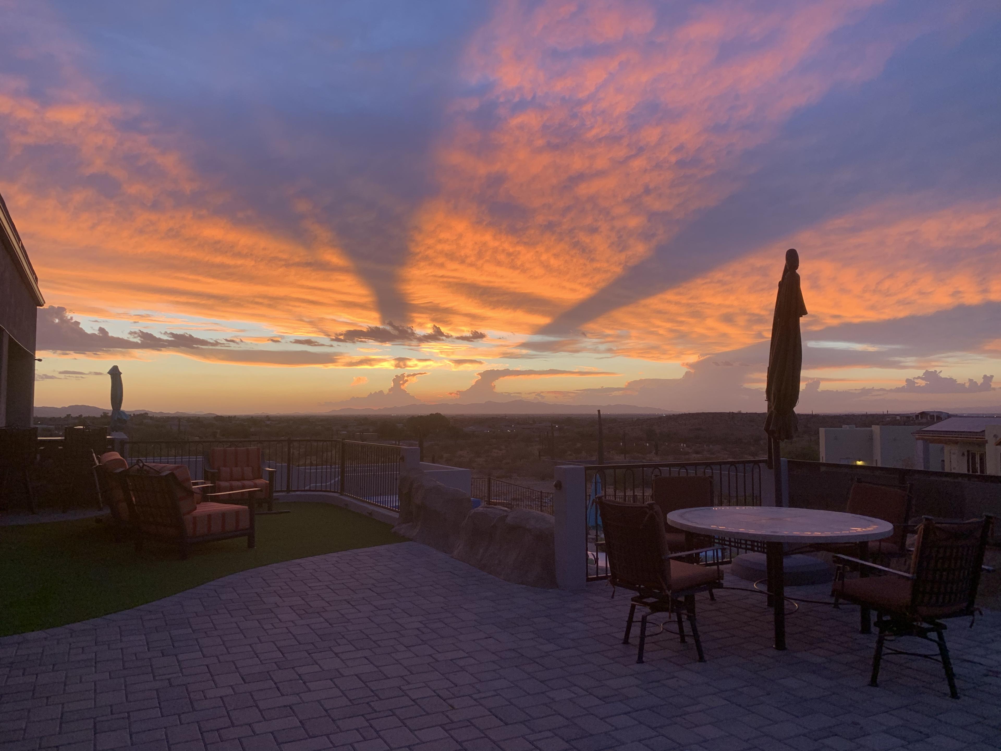 The scene shows a patio with furniture overlooking a vibrant sunset with long shadows stretching across the sky.
