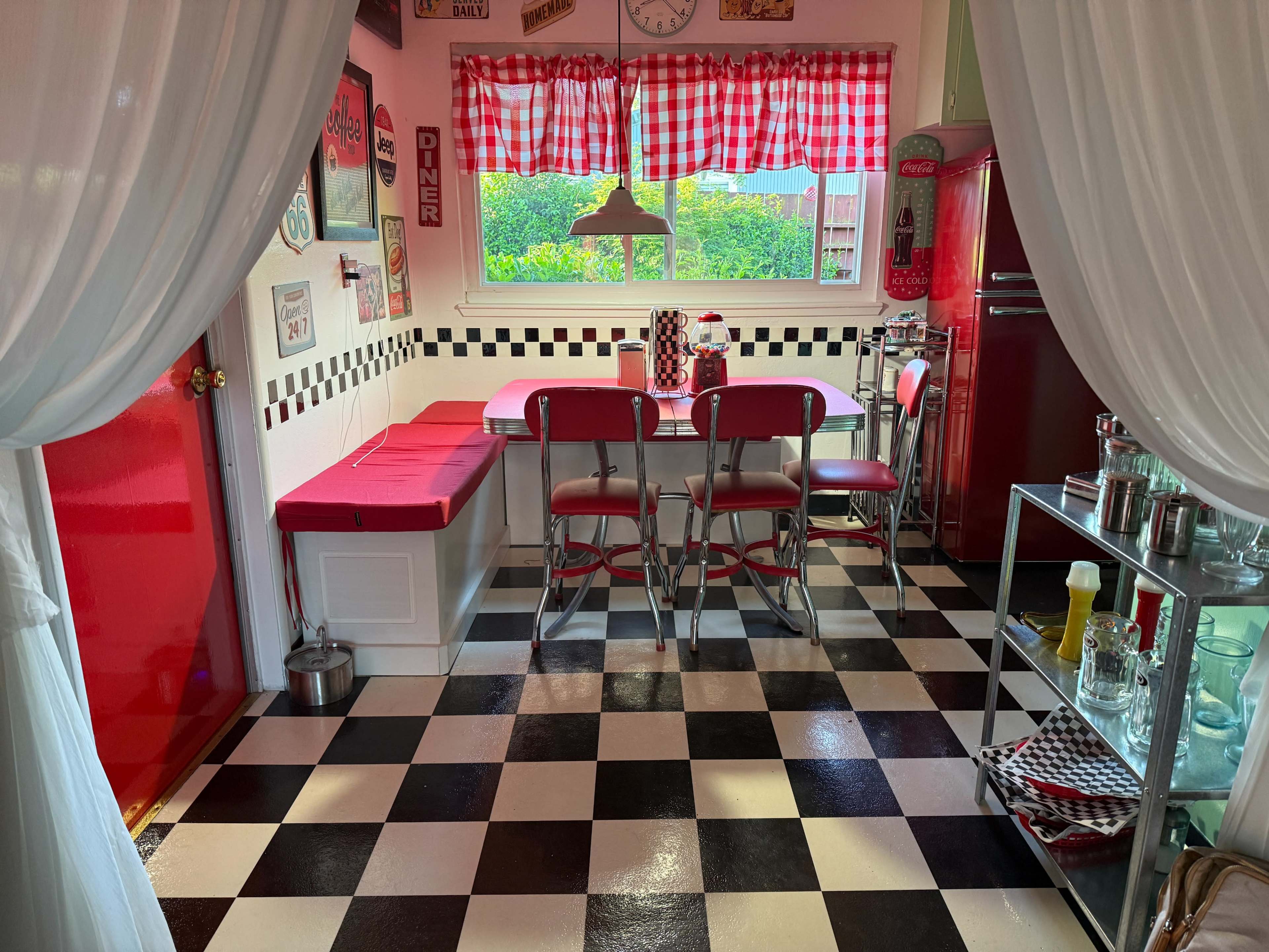 The kitchen features a checkered black and white floor, a red vintage refrigerator, a pink diner-style table with matching chairs, and a window with checkered curtains.