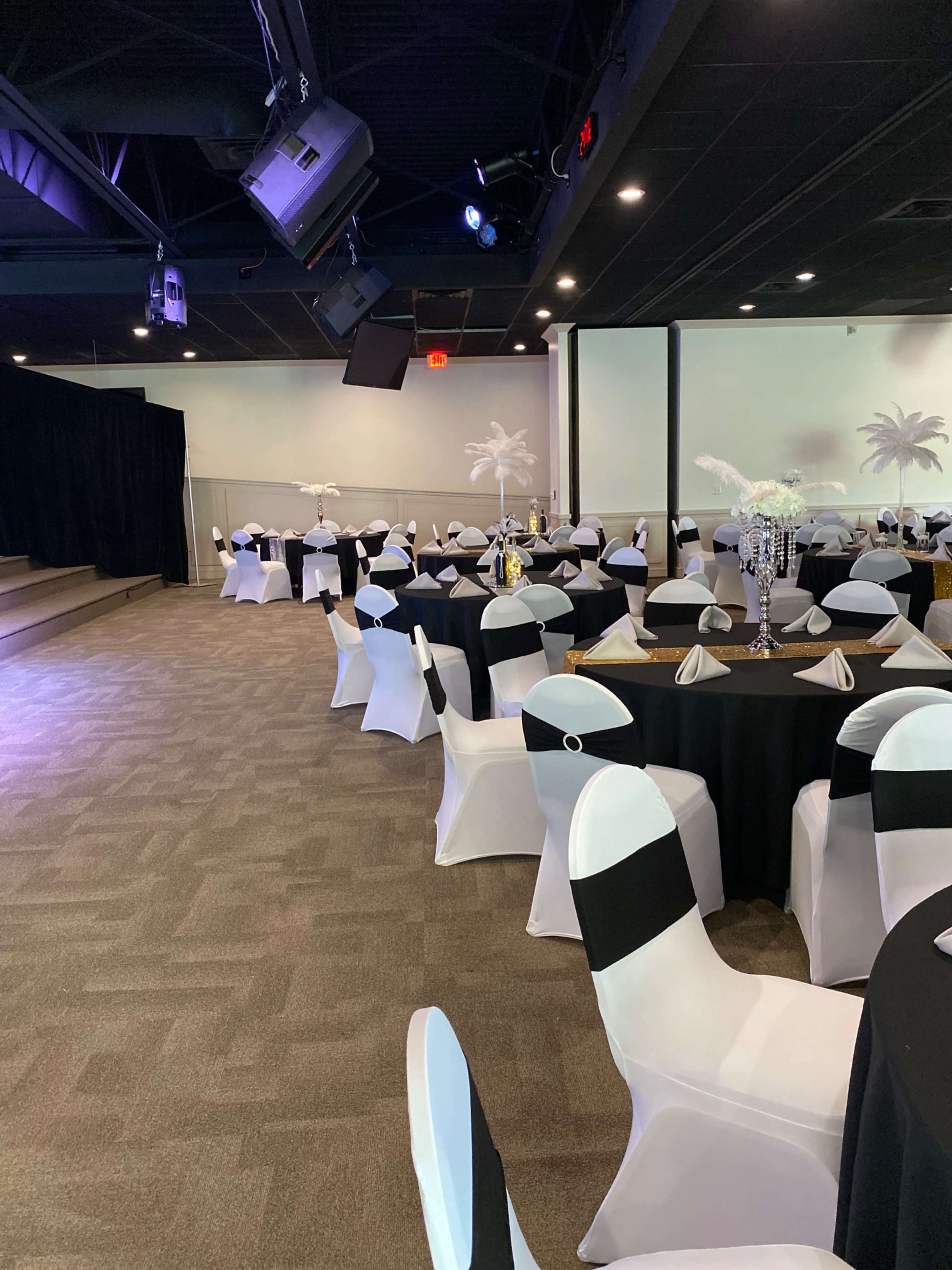 The image shows a banquet hall set up with round tables covered in black and white tablecloths, surrounded by chairs draped in white fabric with black accents.