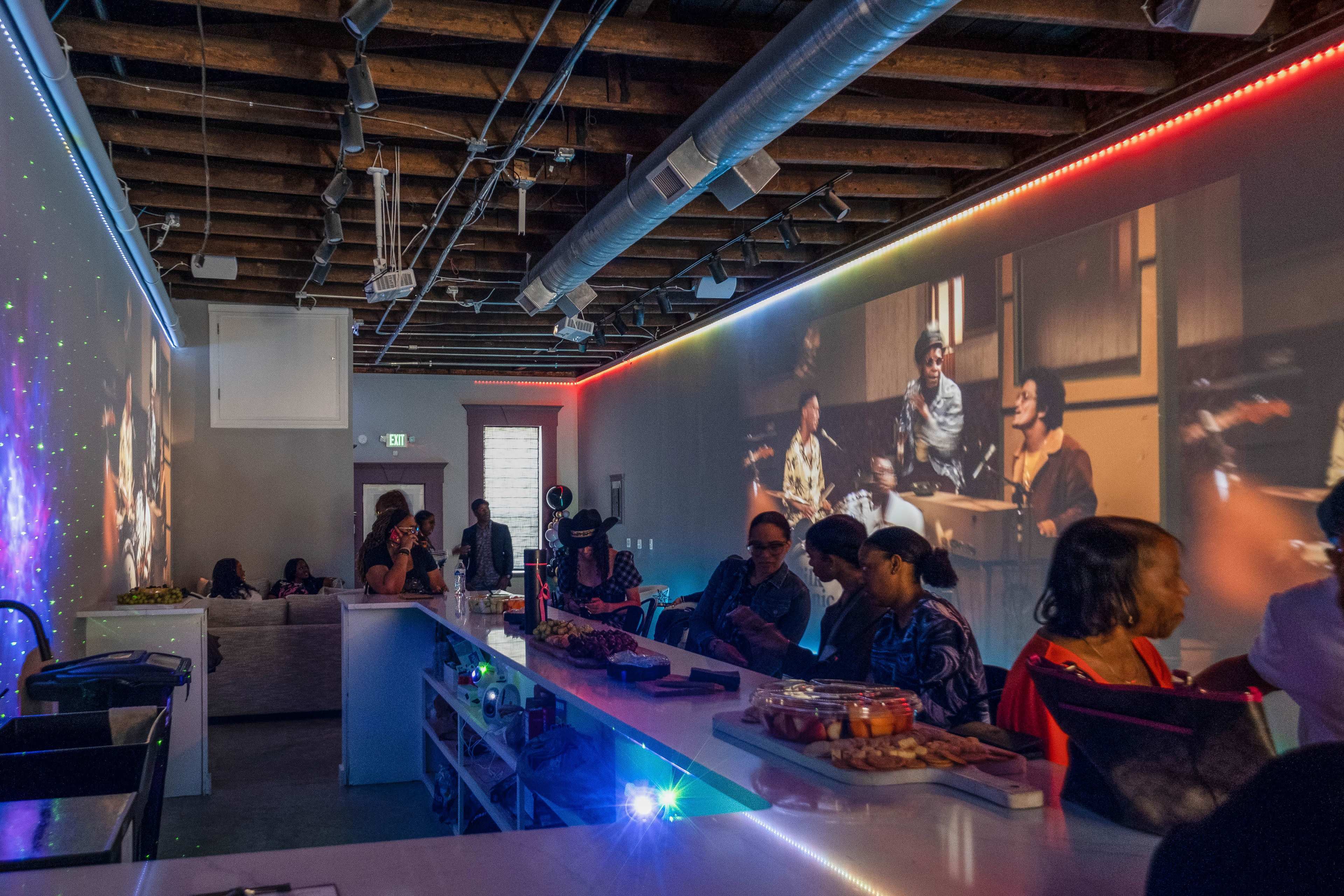 A crowded bar features a long counter with drinks and food, while colorful projections illuminate the walls, showcasing scenes of people interacting.