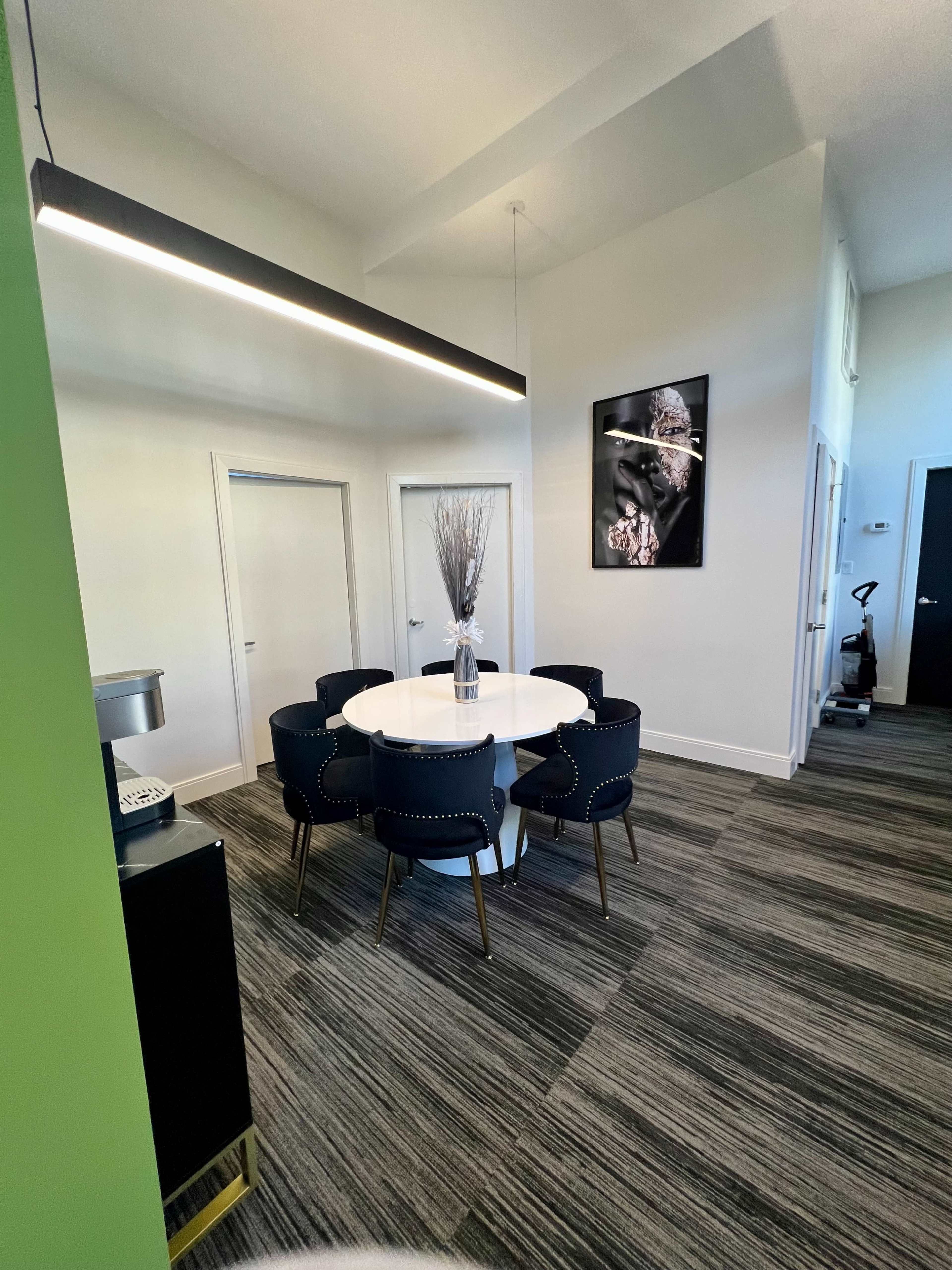 The image shows a modern office space featuring a round table surrounded by six chairs, with a vase of branches on the table and a large framed artwork on the wall.