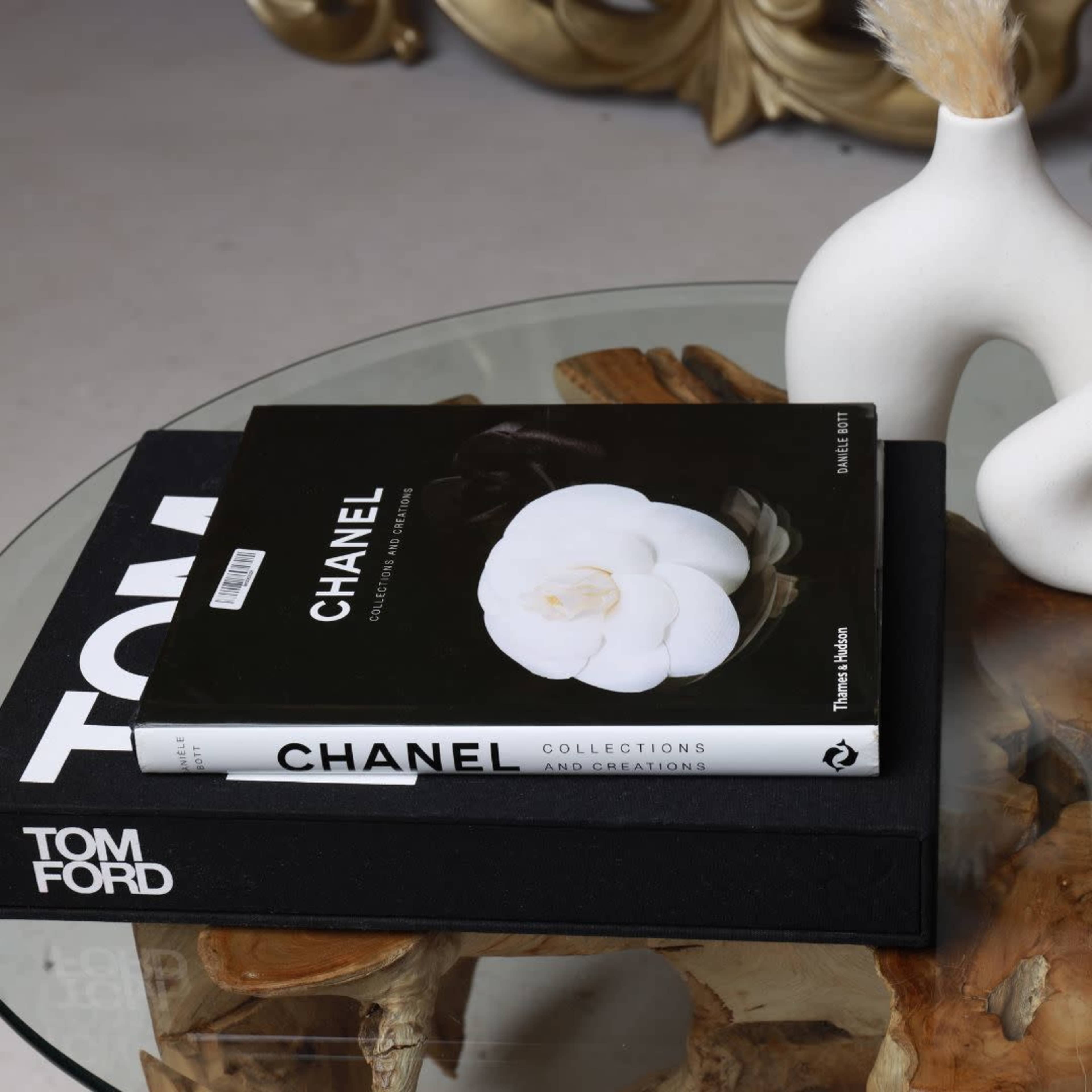 A stack of stylish coffee table books, including "Chanel" and "Tom Ford," sits on a glass table with a decorative vase and dried plant.