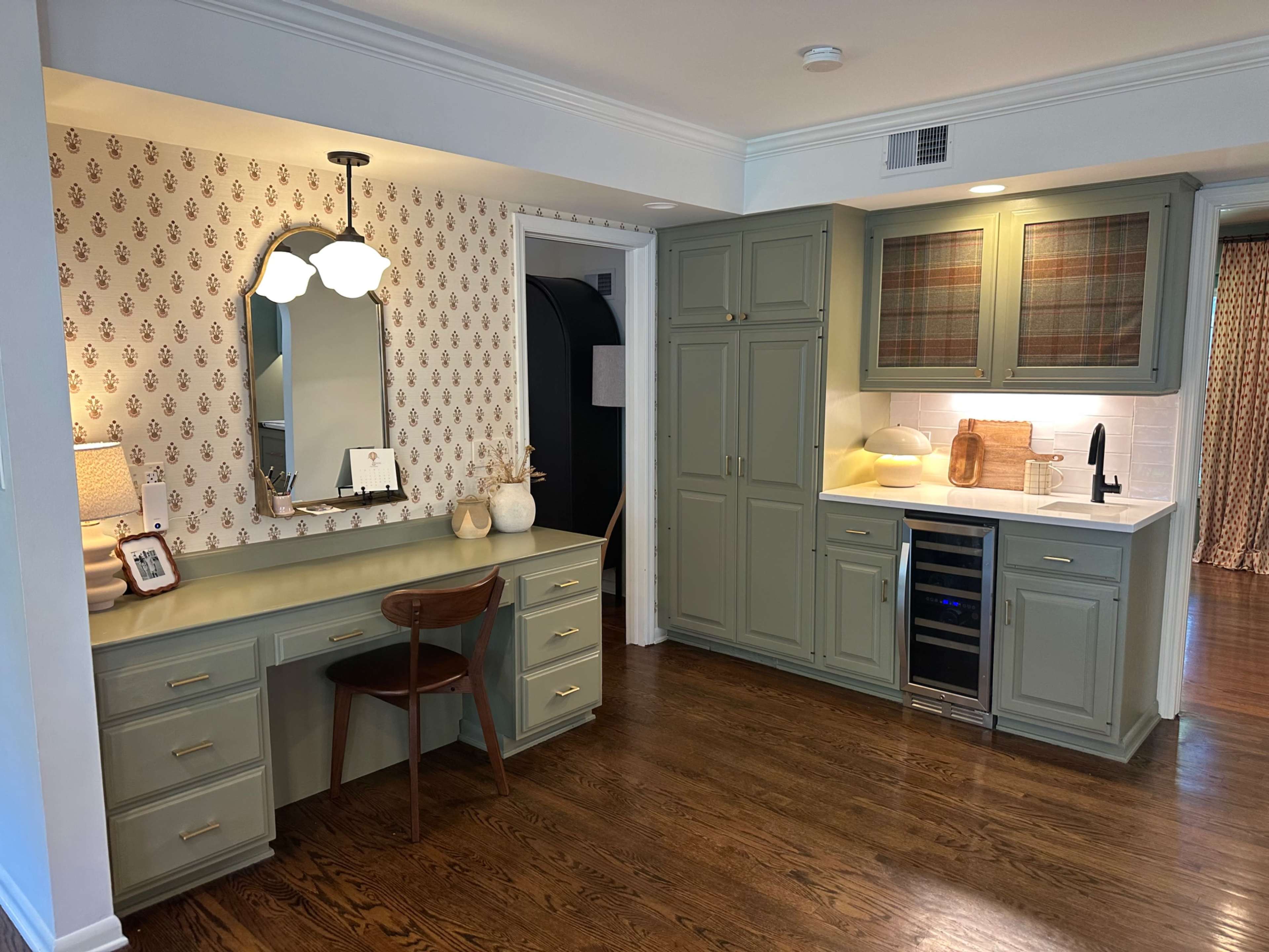 The image shows a cozy kitchen area with a light green cabinetry, a countertop with a wine cooler, and a matching desk workspace beside a patterned wall.