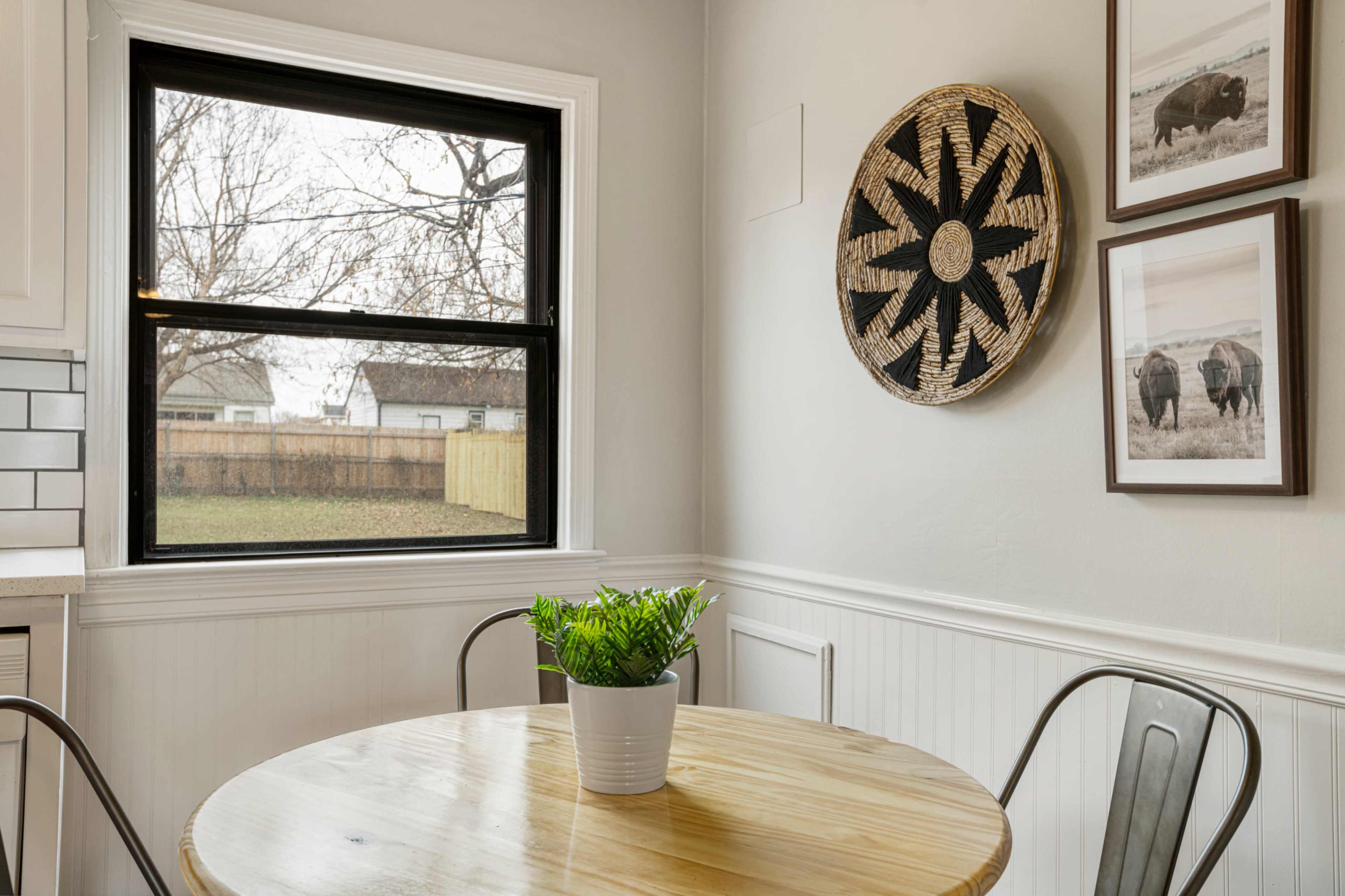 A circular wooden table with metal chairs is positioned next to a window, featuring decorative wall art and a small potted plant above a white wainscoted wall.