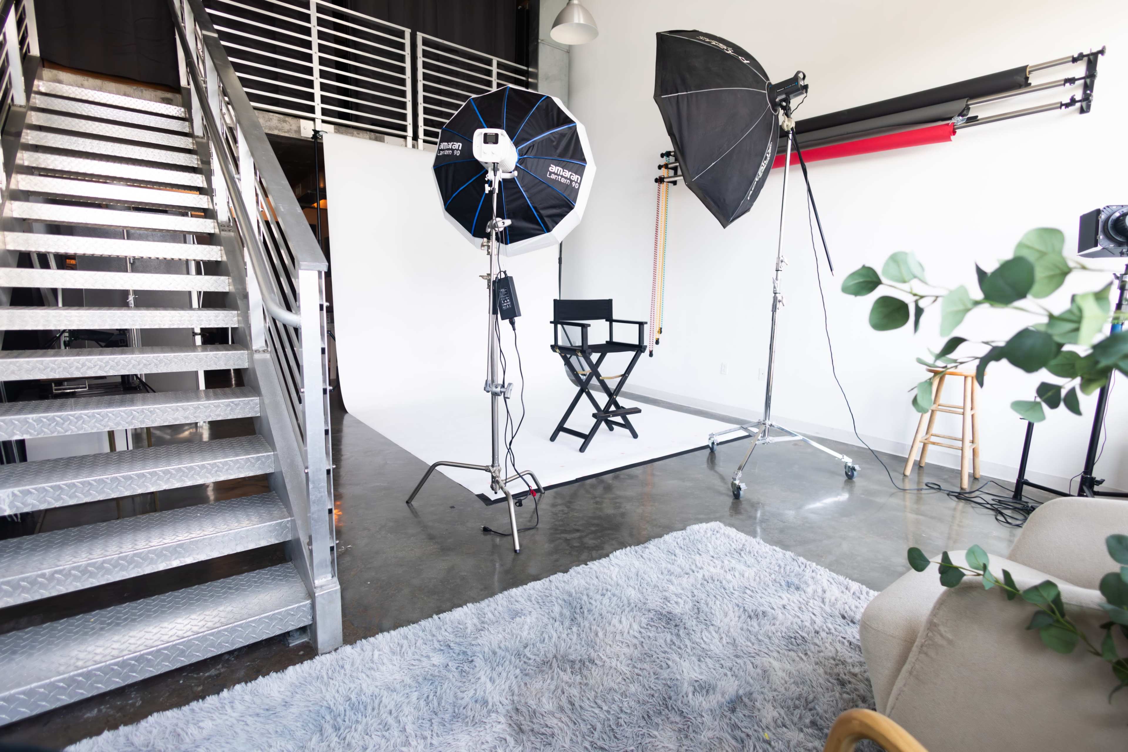 The image shows a photography studio with a chair, two large softbox lights, a staircase, and a rug on the floor.