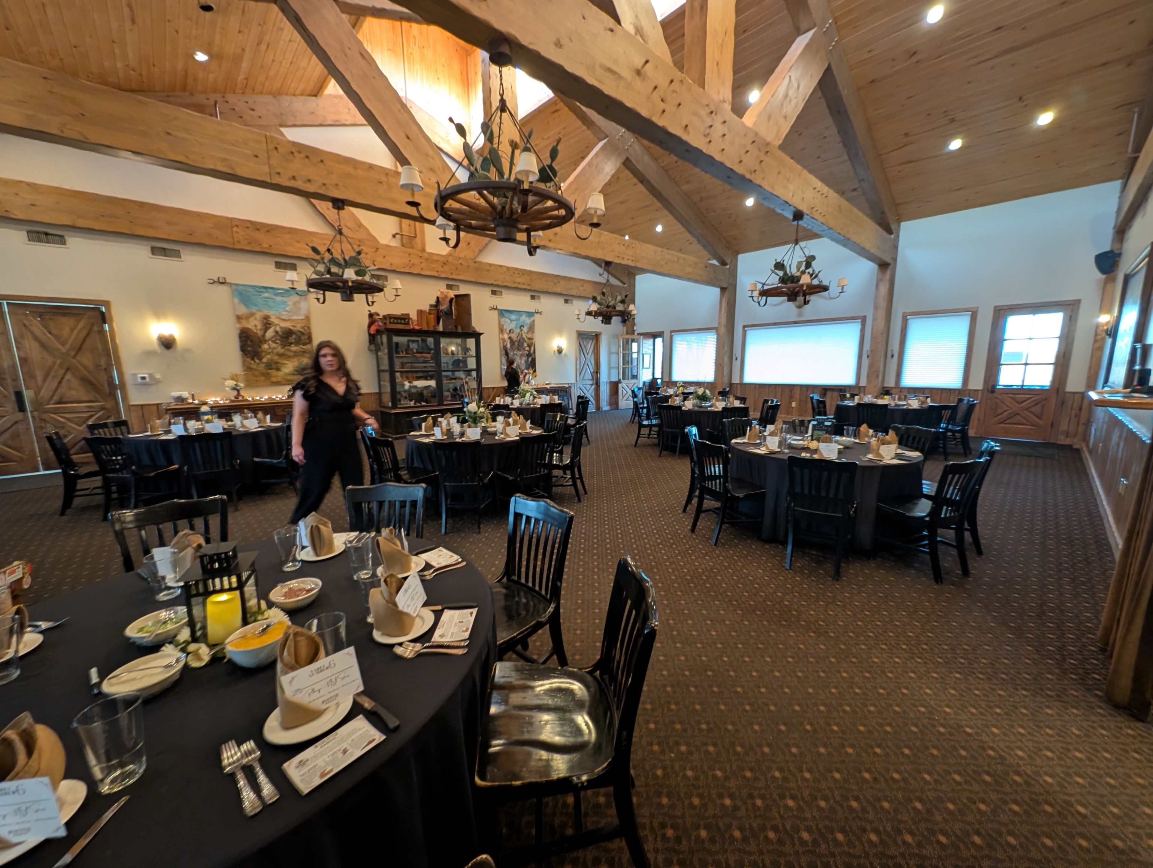 The image shows a spacious dining room with wooden beams, neatly arranged round tables set for a meal, and a staff member walking toward the camera.