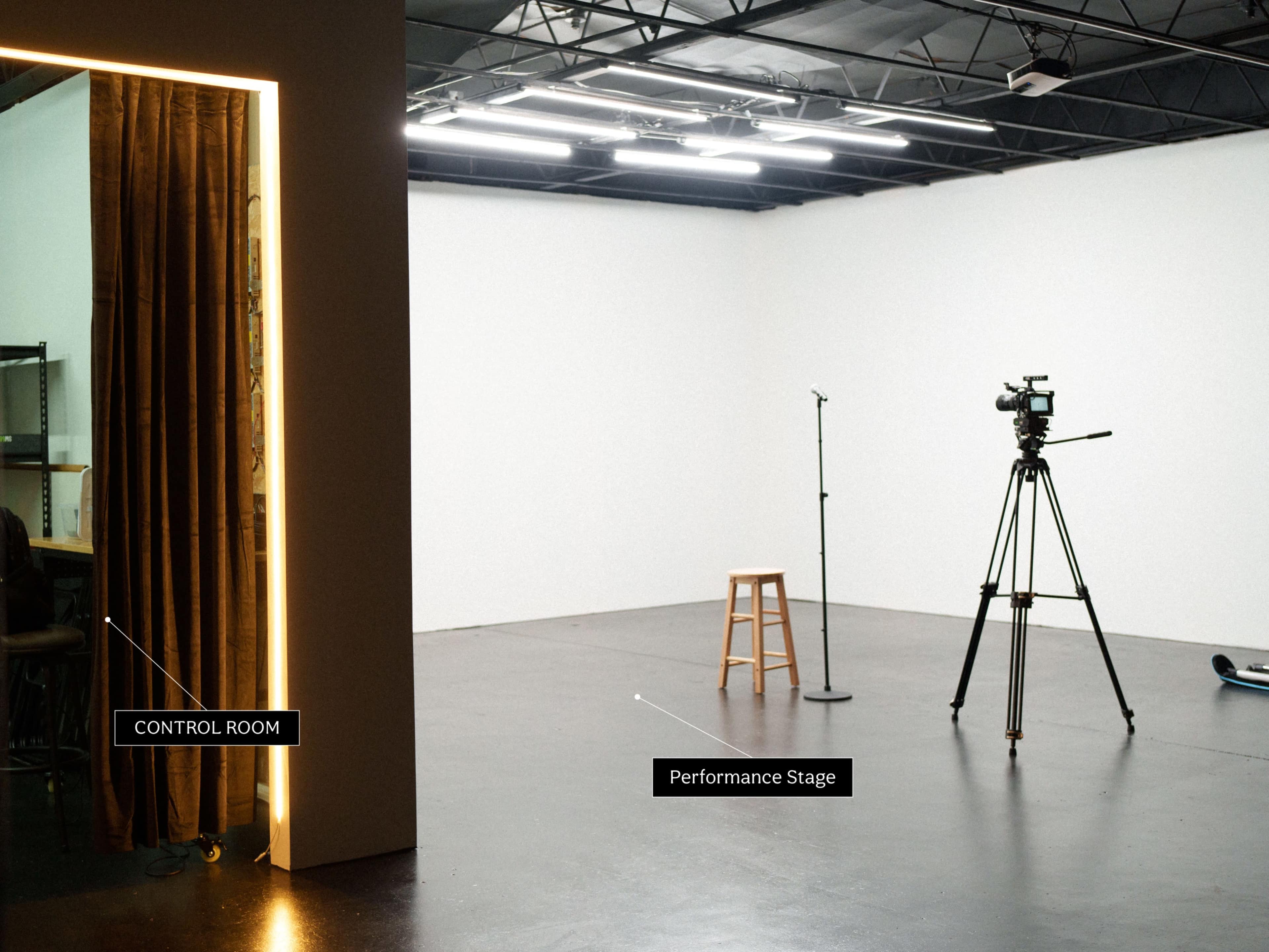 The image shows a minimalist studio space featuring a performance stage with a microphone and a stool, adjacent to a control room separated by a curtain.