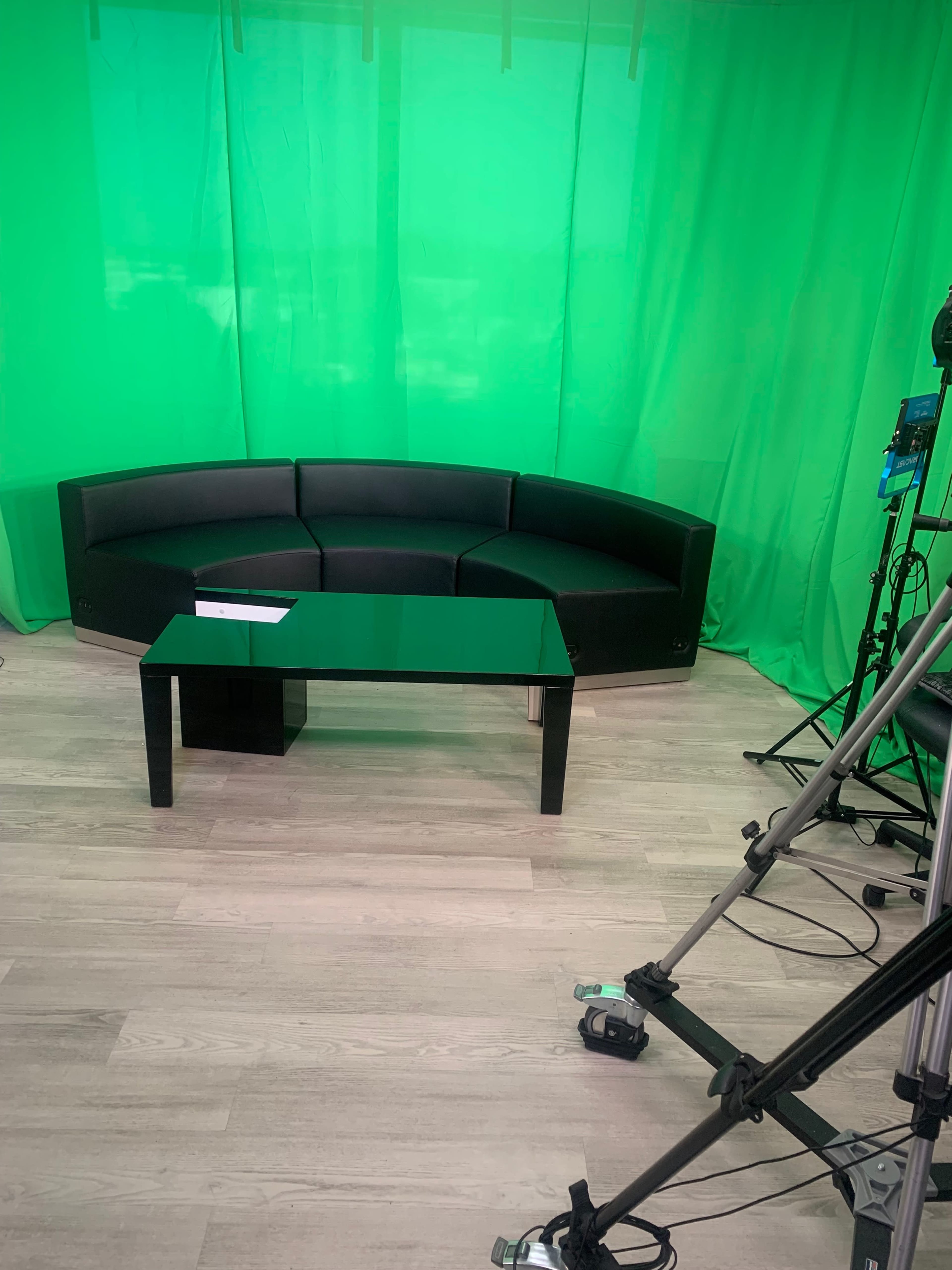 The image shows a black curved sofa and a black table in front of a green backdrop, with a tripod and camera equipment to the side.