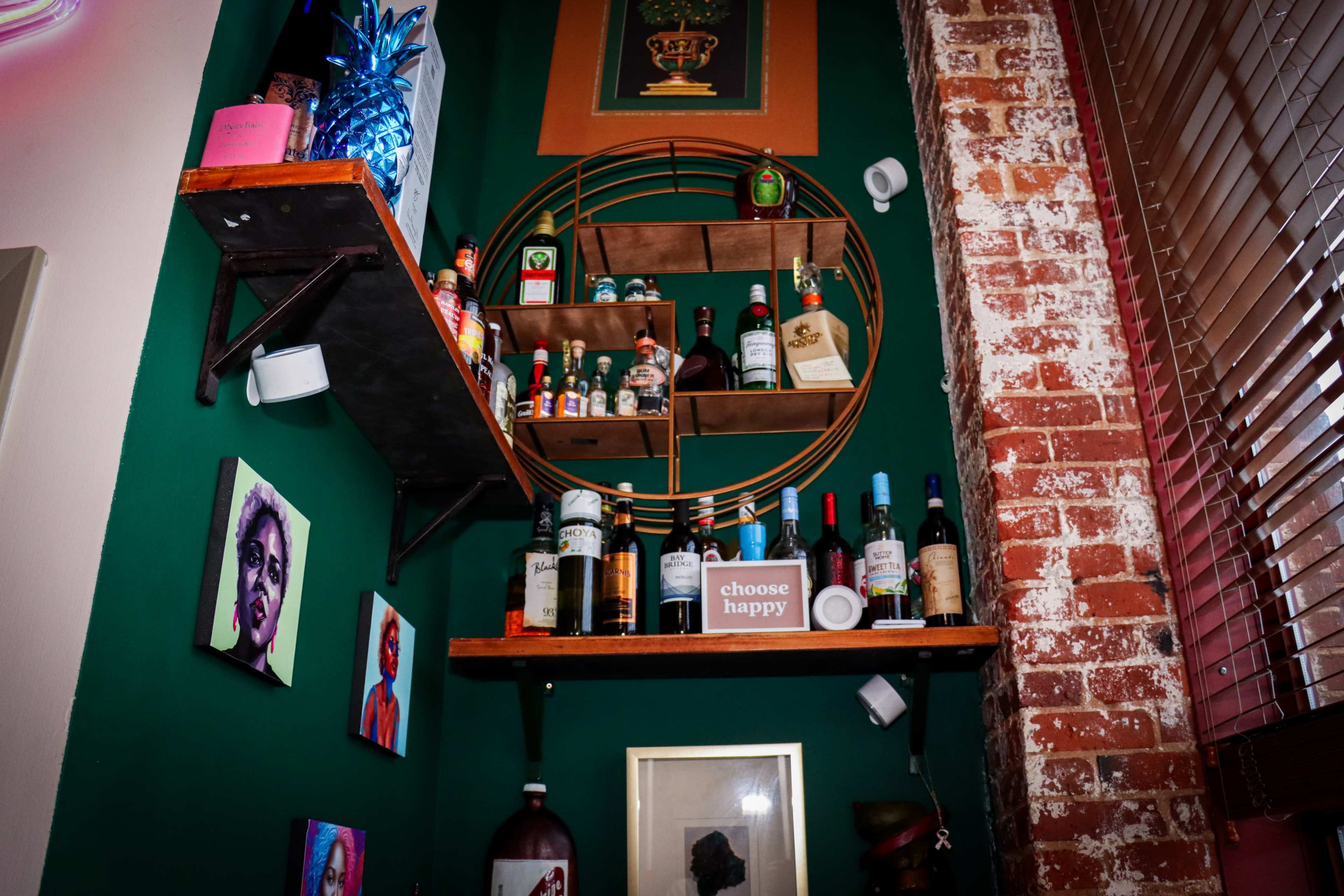 A corner of a room with green walls displaying various bottles of drinks on wooden shelves, alongside framed artwork and a wall adorned with exposed brick.
