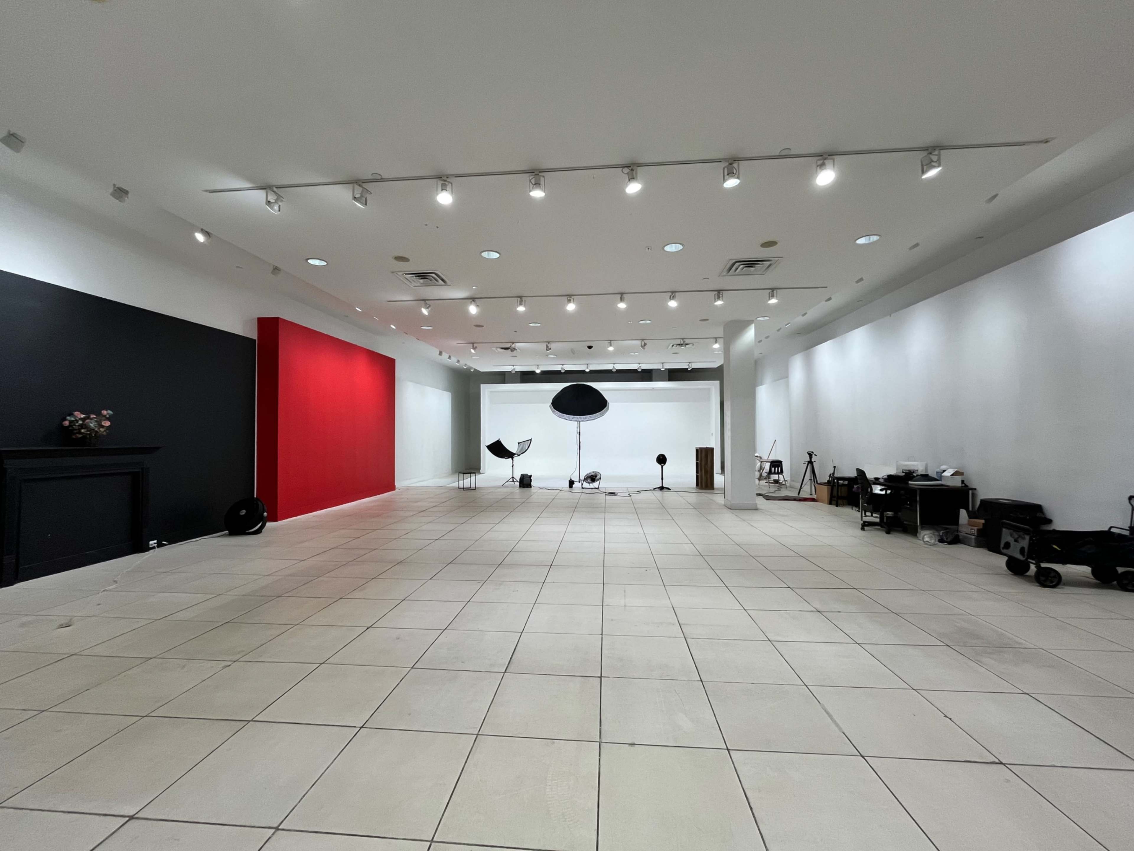 The image shows a spacious studio or gallery with a white interior, featuring a large red wall, a black wall, and various photography equipment arranged throughout the room.