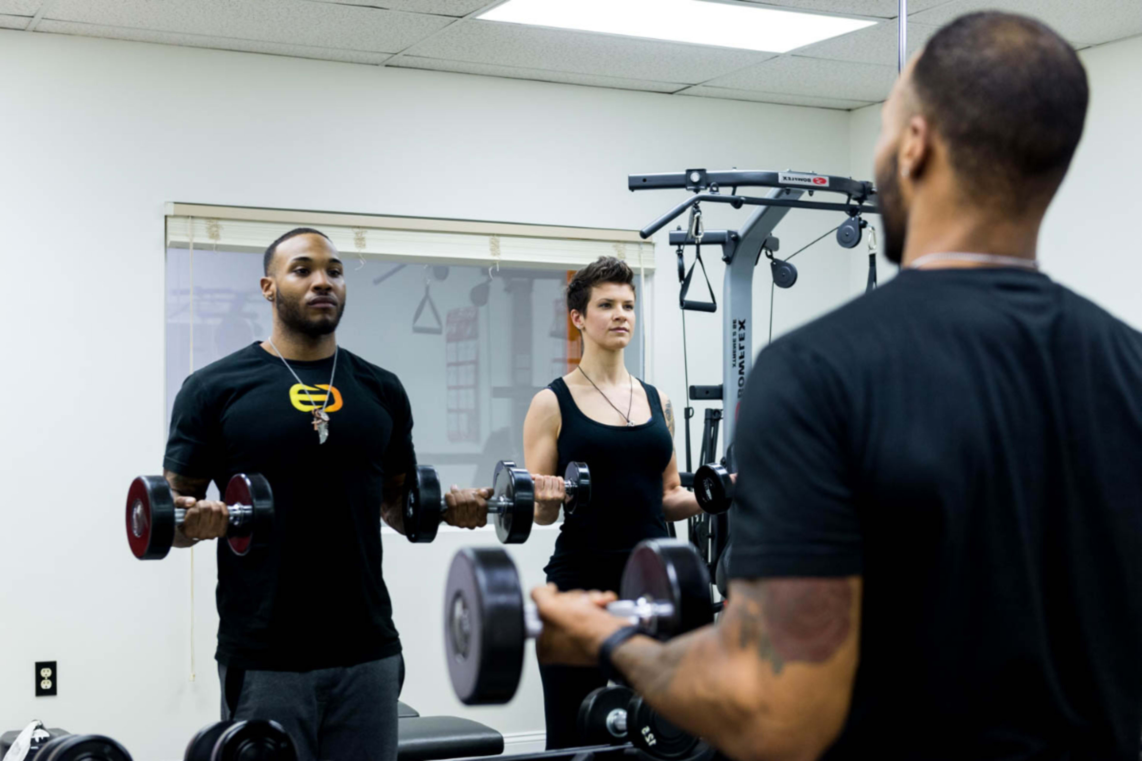 A man and a woman are exercising in a gym, each holding dumbbells while facing a mirror.