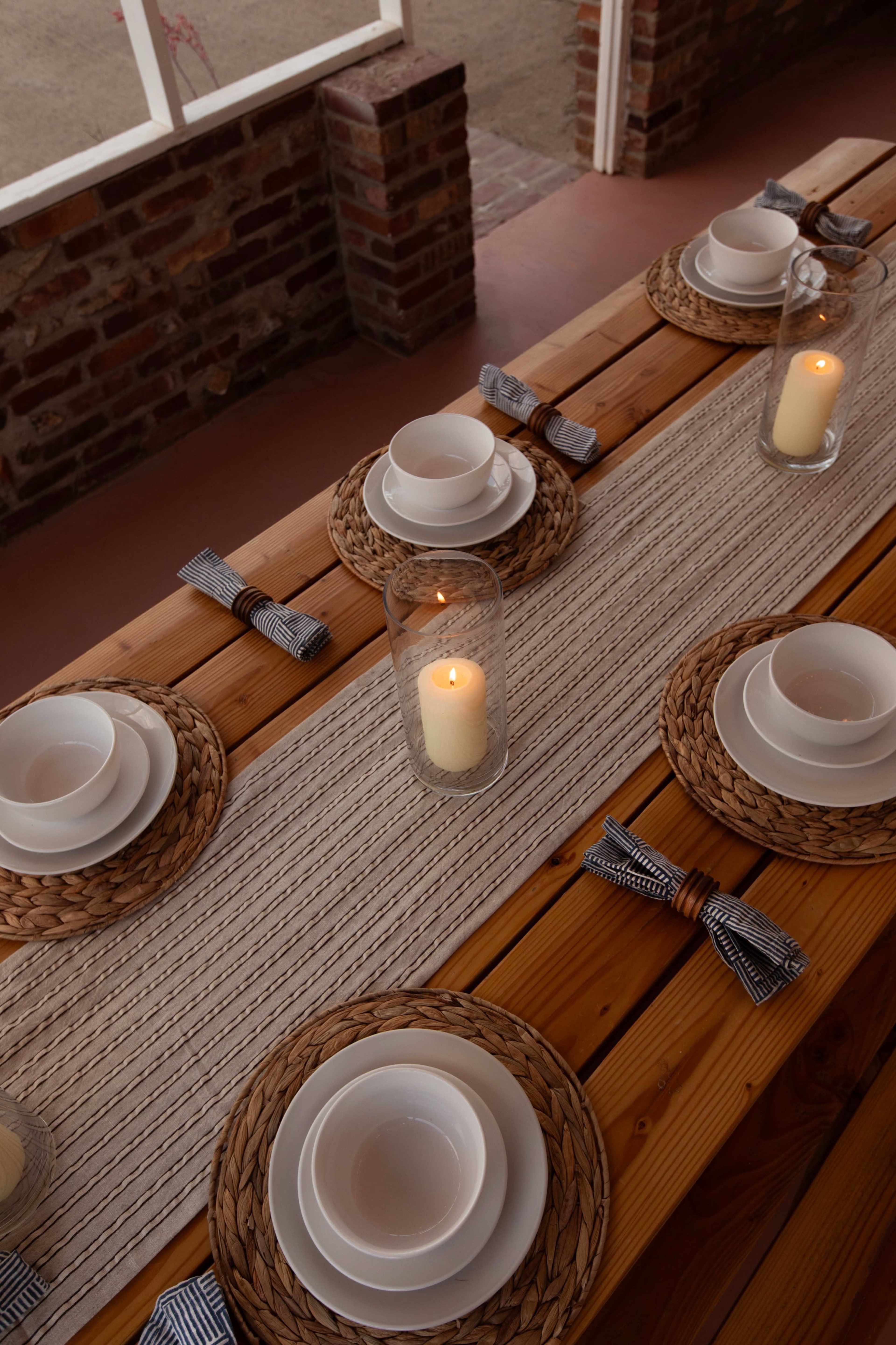 A wooden dining table is set with woven placemats, white bowls, and candles arranged along a textured runner.