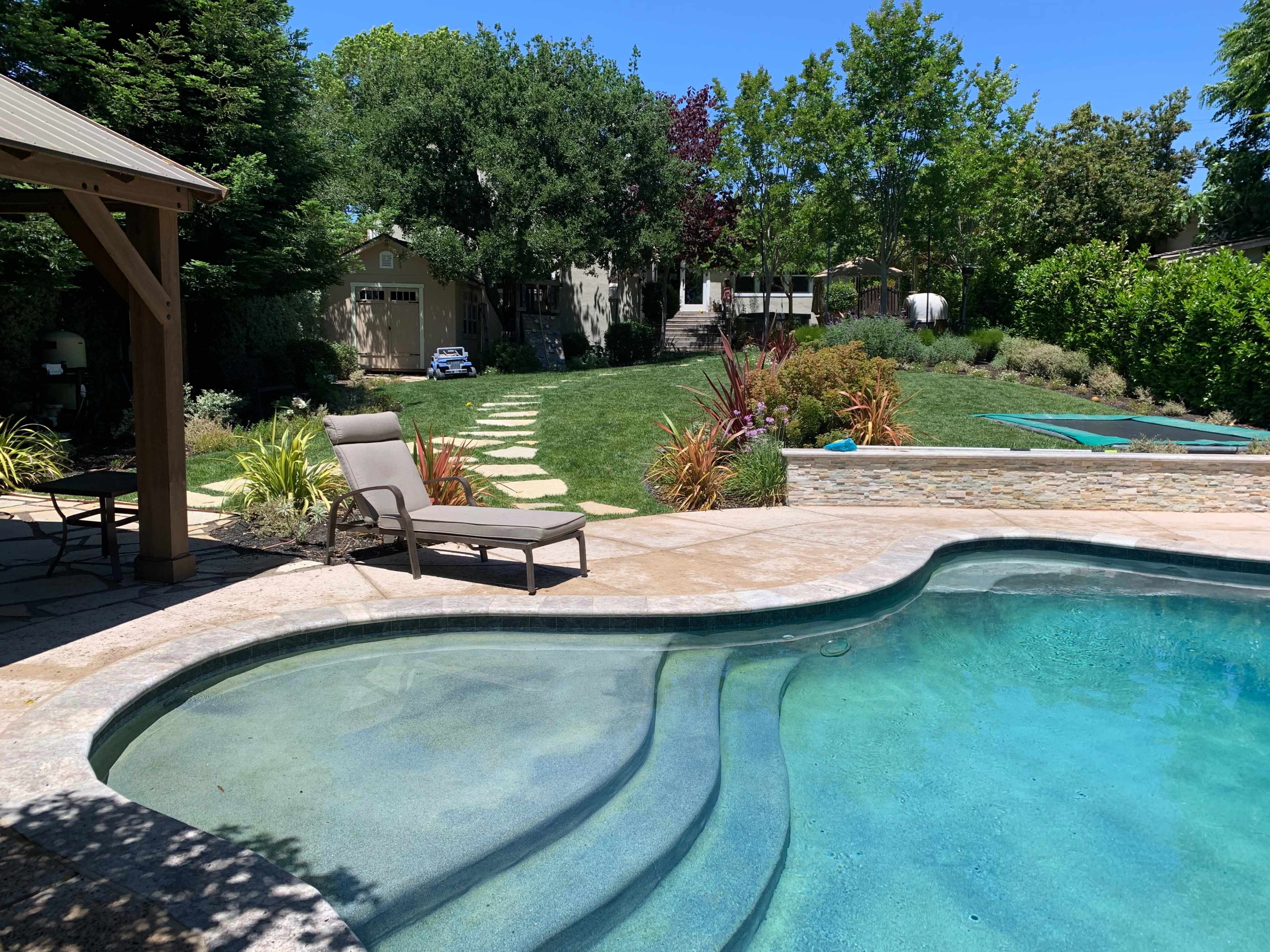 A swimming pool is situated in a backyard with a stone patio, surrounded by landscaping and a grassy area leading to a pathway and a house in the background.